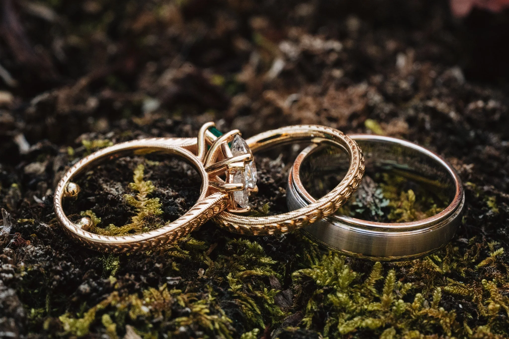 Three rings, one with a diamond and emerald accents, placed on moss and soil.