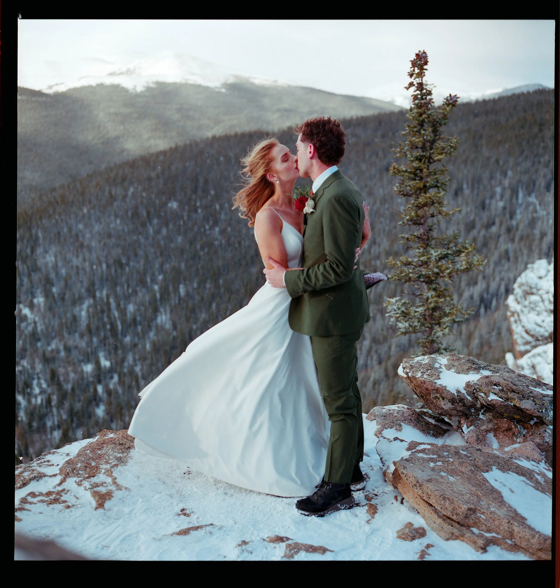 A couple in wedding attire sharing a kiss on a snowy mountain ledge with a backdrop of forested mountains and snow-capped peaks.