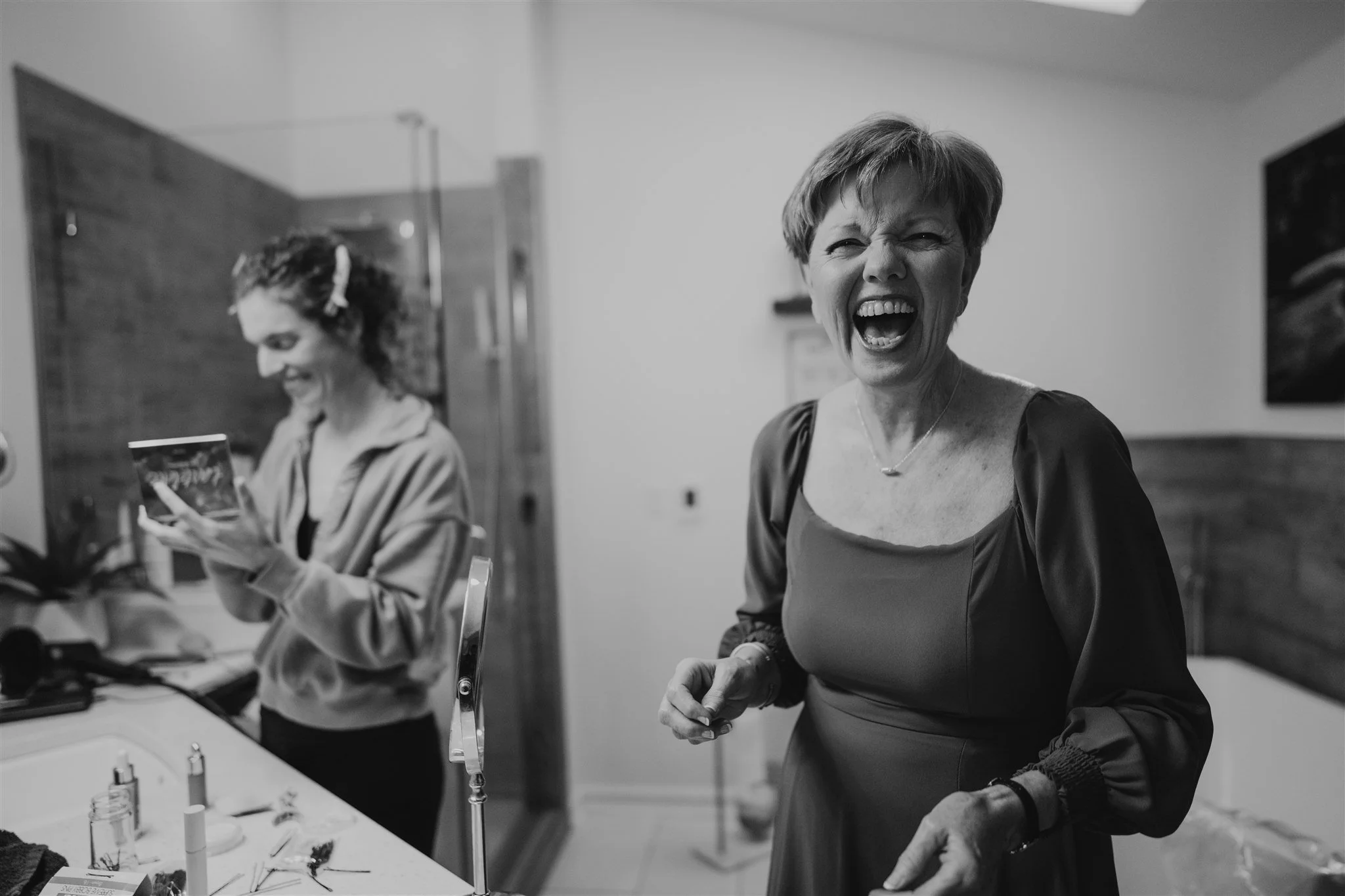 Two women laughing and smiling in a room, one with short hair wearing a dress, the other with curly hair wearing a hoodie, surrounded by various objects on a table.