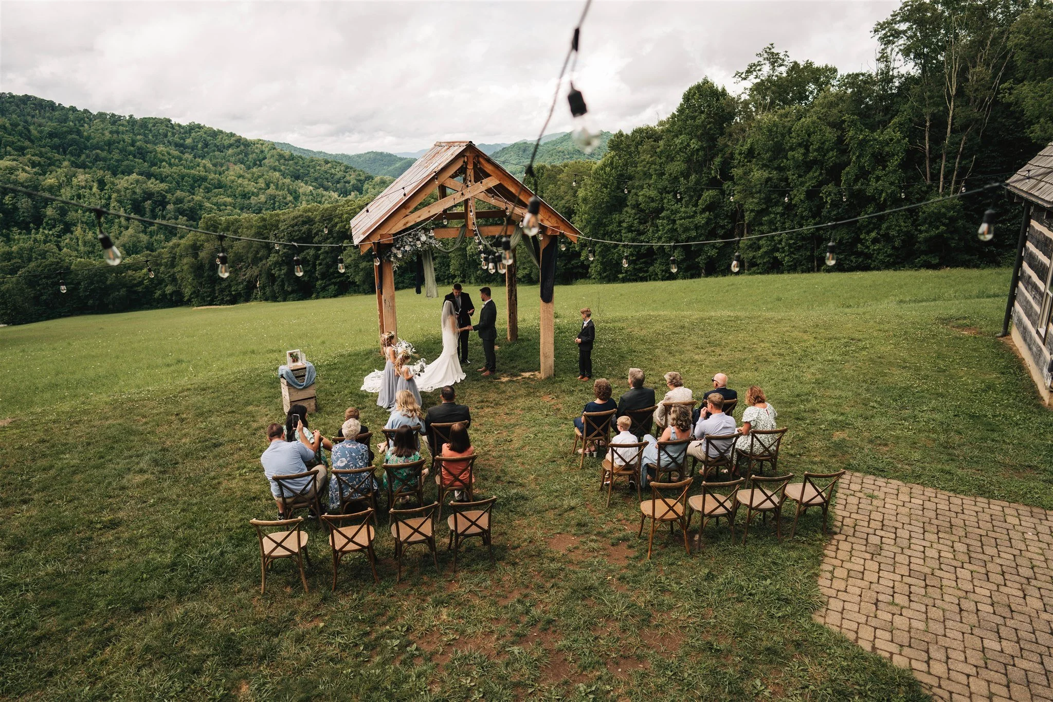 Outdoor wedding ceremony taking place on a grassy field with mountains in the background. The bride and groom are standing under a wooden pergola with floral decorations, exchanging vows. Guests are seated on chairs facing the couple, with string lights hanging overhead.