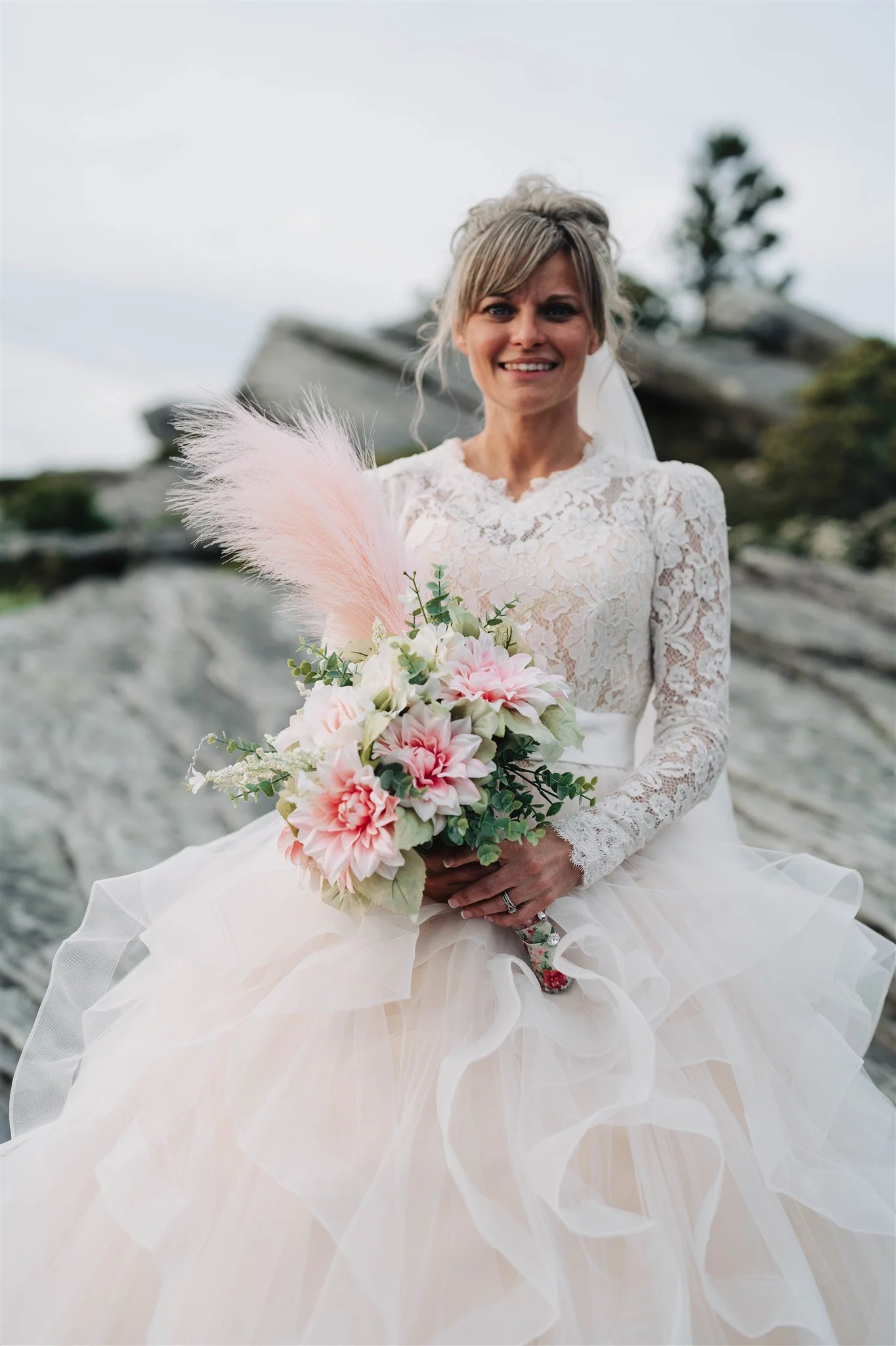 A bride in a lace wedding dress sitting outdoors on rocks, holding a bouquet of pink and white flowers with greenery and pink pampas grass.