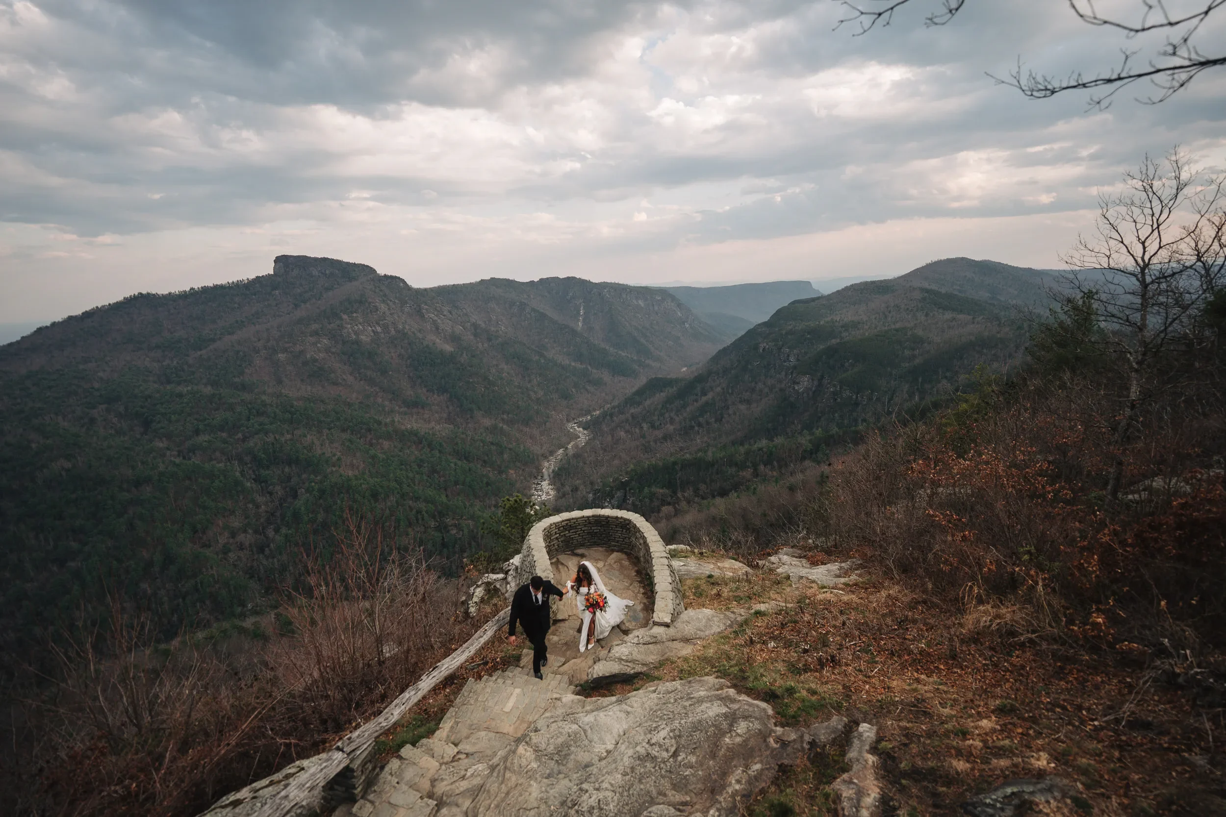 Couple walking up stone steps toward a mountain overlook with sweeping gorge views and dramatic clouds overhead.