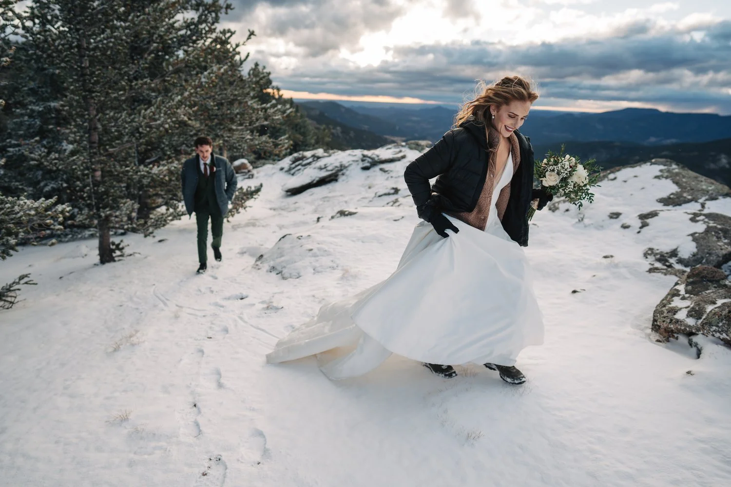 Woman in wedding dress holding bouquet, walking in snow on mountain, man in suit following behind.