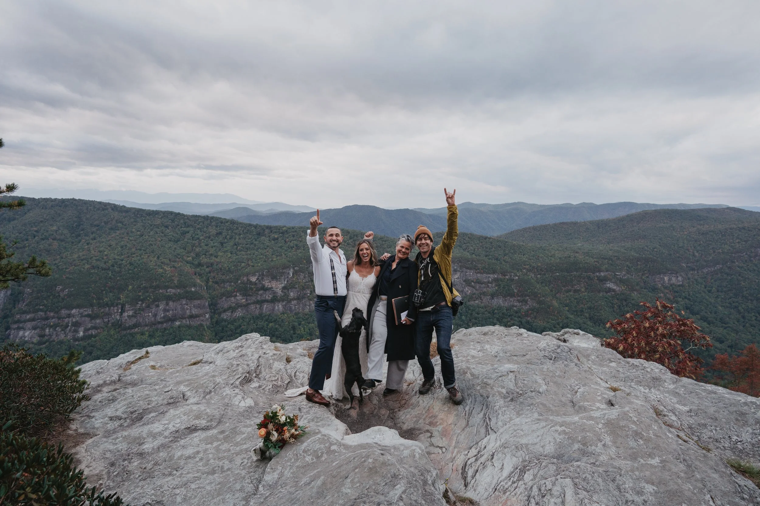 Group portrait on a cliff overlook: elopement couple with their officiant and photographer, smiling with sweeping mountain views behind.