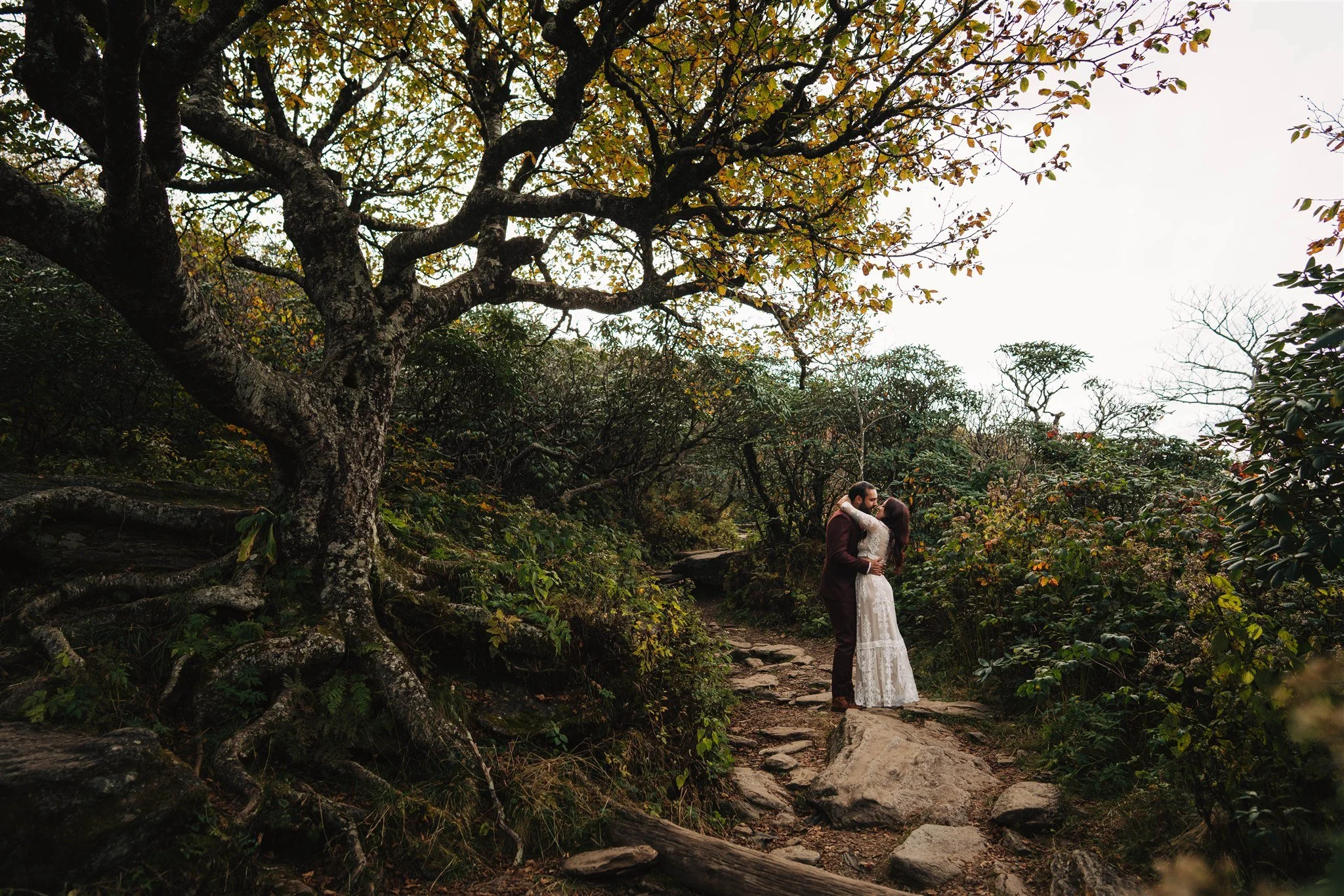 A couple in wedding attire embrace on a rocky forest trail surrounded by trees with sparse autumn foliage.