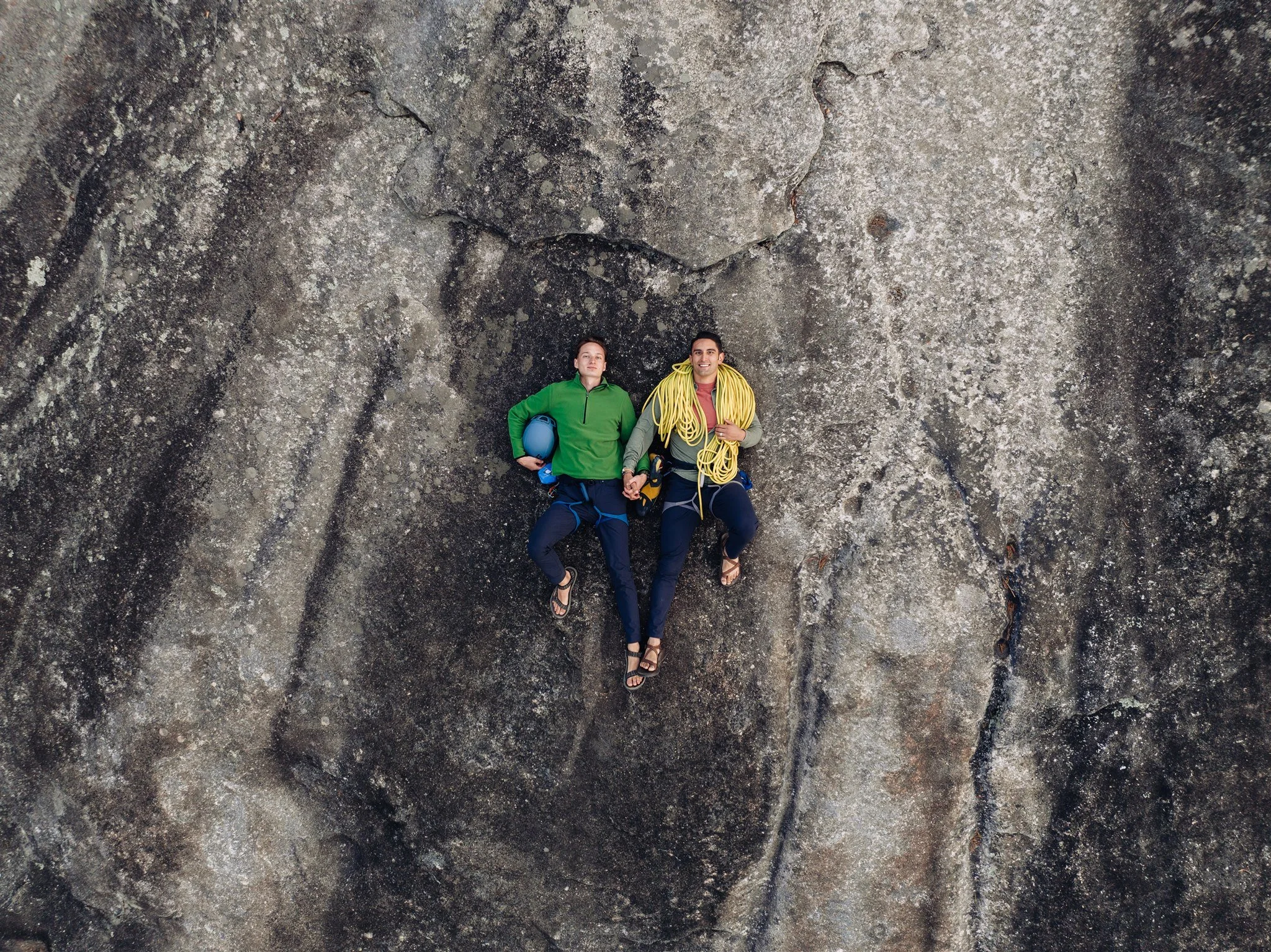 Two rock climbers lying on a rock face, one holding climbing gear and a rope, wearing outdoor clothing.