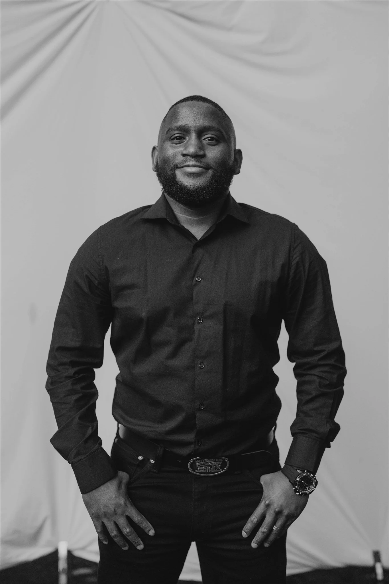 A confident man wearing a black long-sleeve shirt, black pants, and a watch standing against a light backdrop.