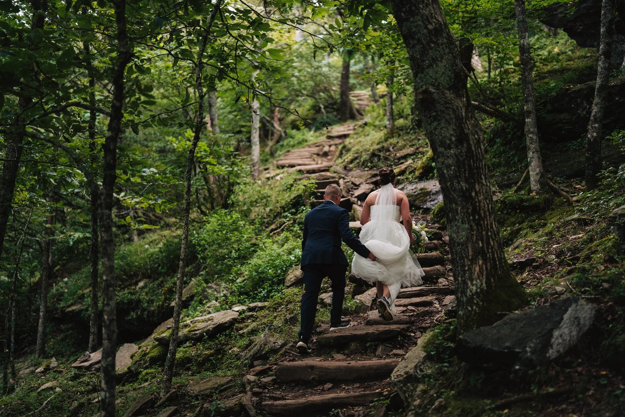 A bride and groom walking up a rocky forest trail, surrounded by green trees and foliage.
