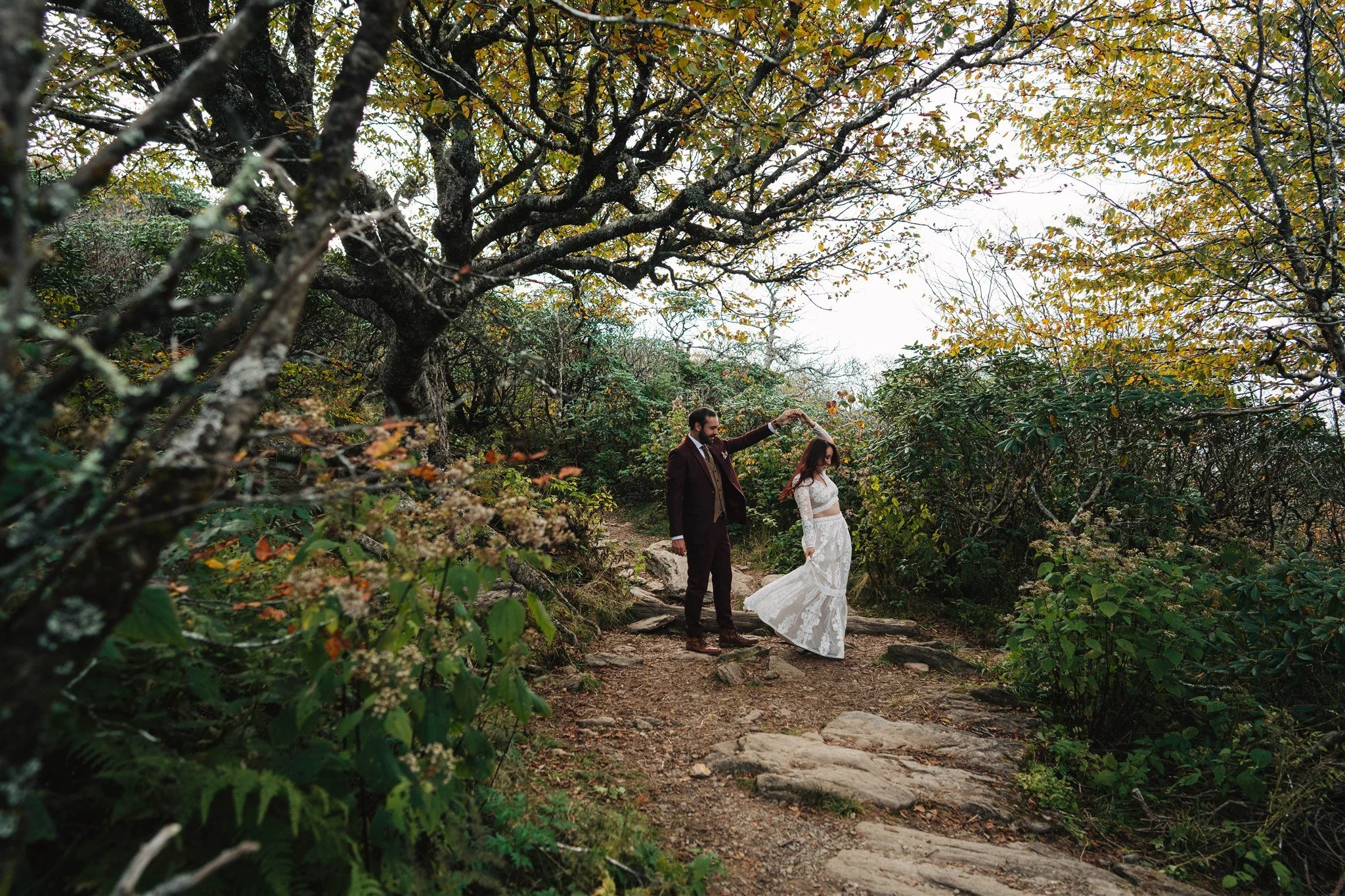 A man and a woman, dressed in formal wedding attire, holding hands as they walk along a narrow dirt trail surrounded by lush green foliage and trees with autumn leaves.