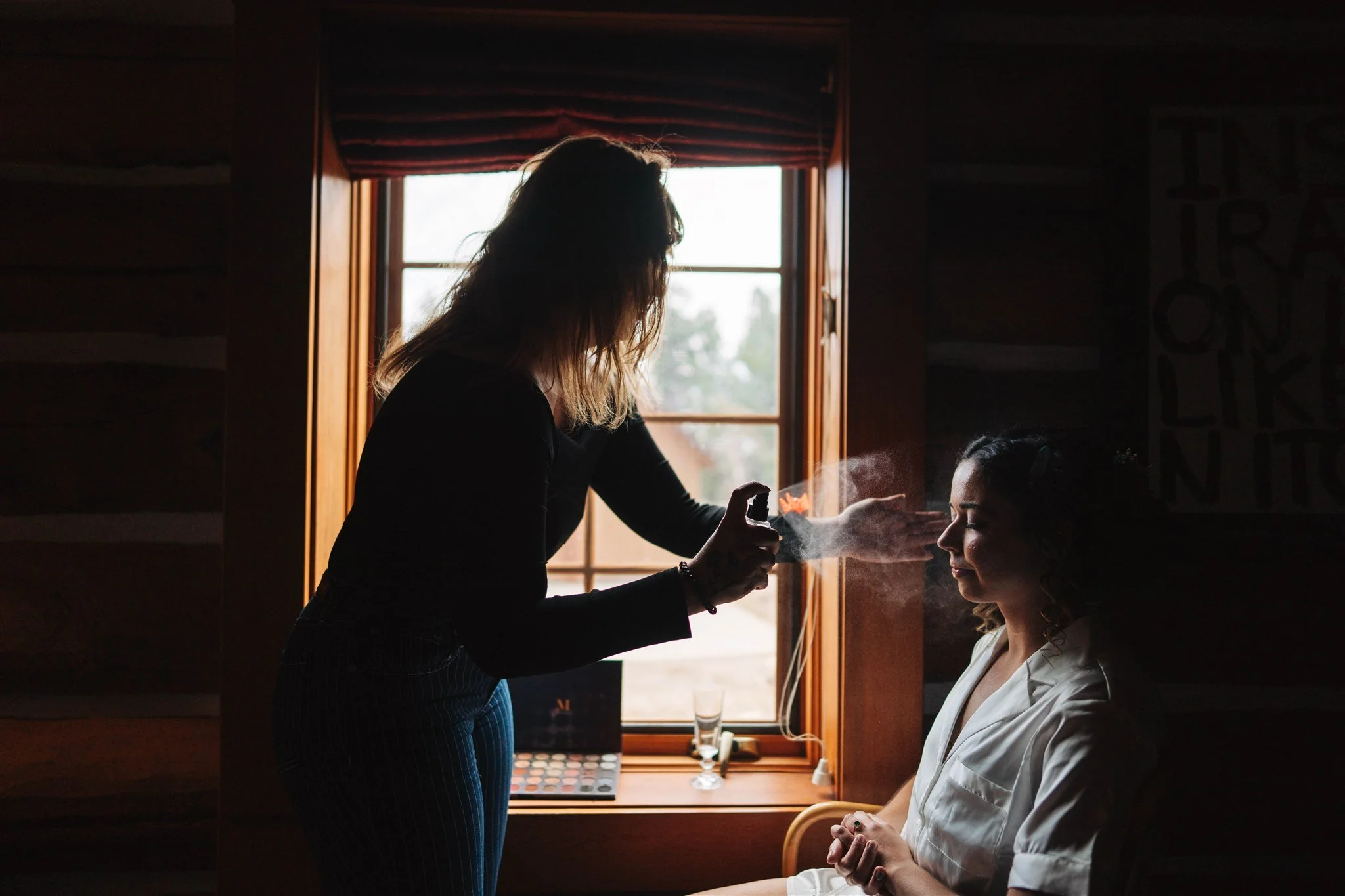 Makeup artist applying setting spray on a woman near a window.