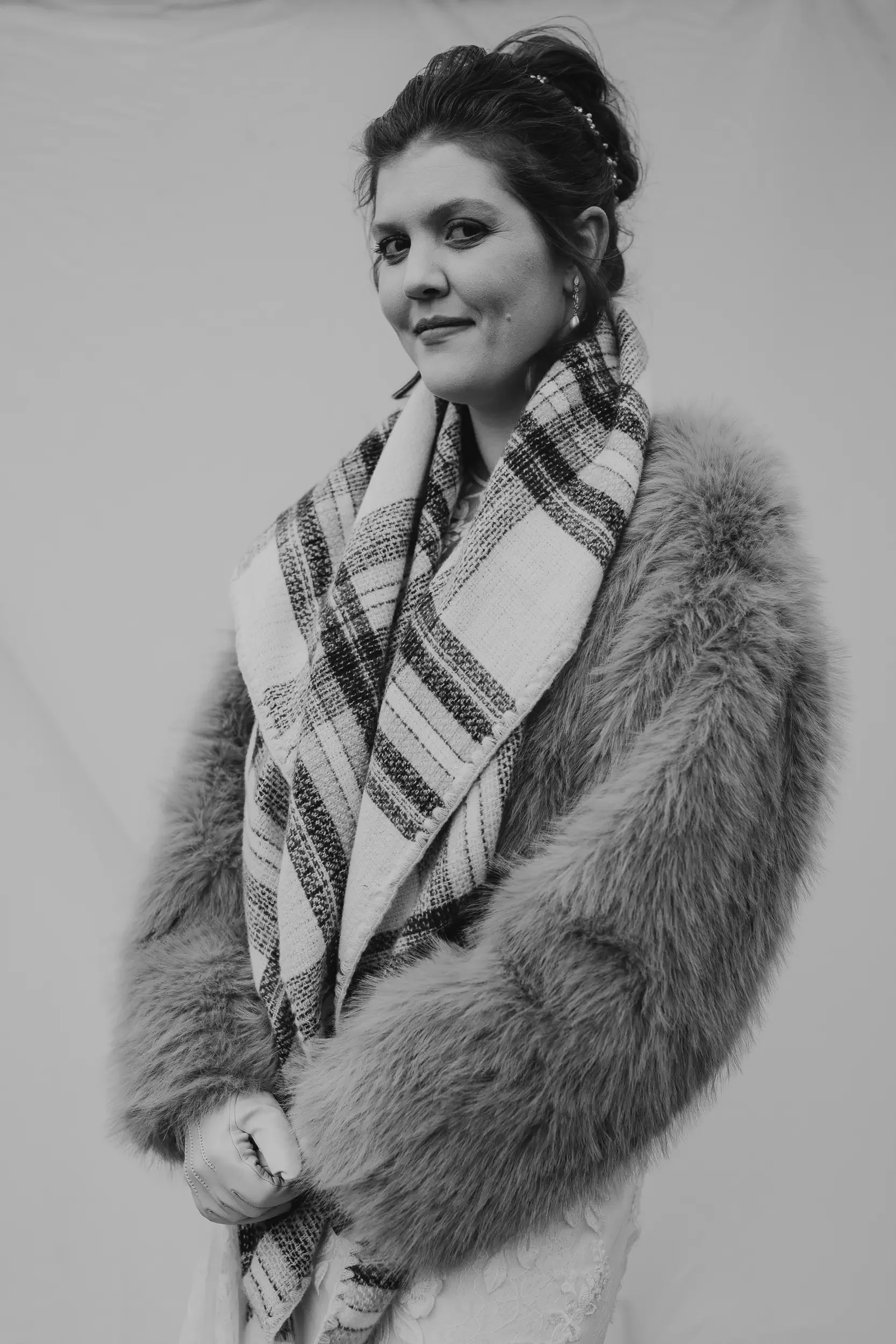 A woman with dark hair styled up, wearing a fur coat and a plaid scarf, standing against a plain background in a black and white photo.