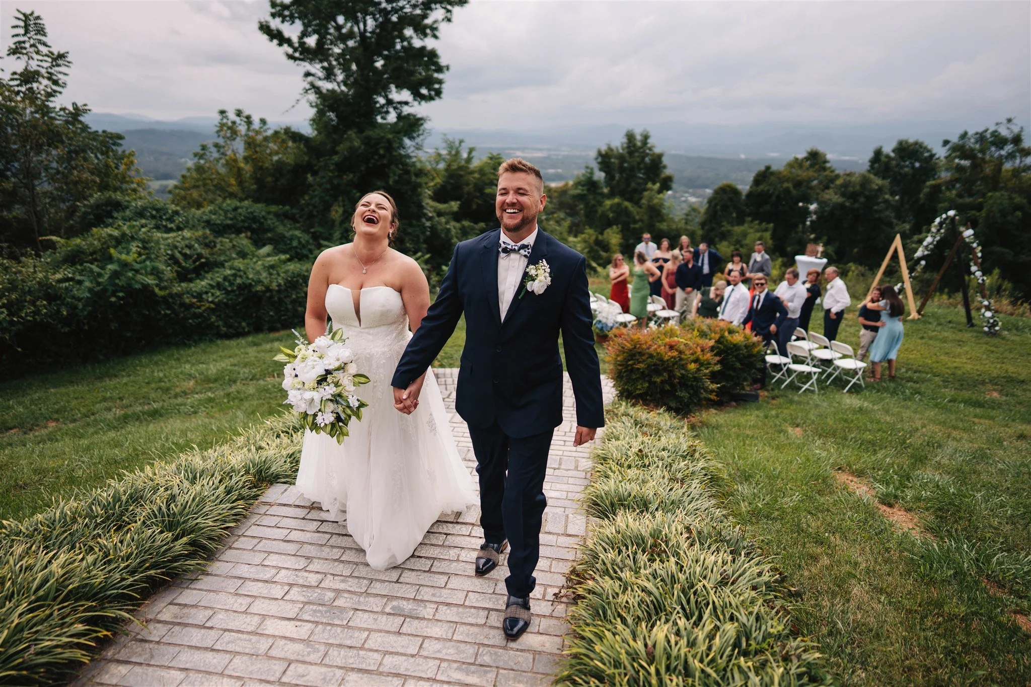 A newlywed couple walking hand in hand outdoors after a wedding ceremony, with guests in the background and scenic mountains in the distance.