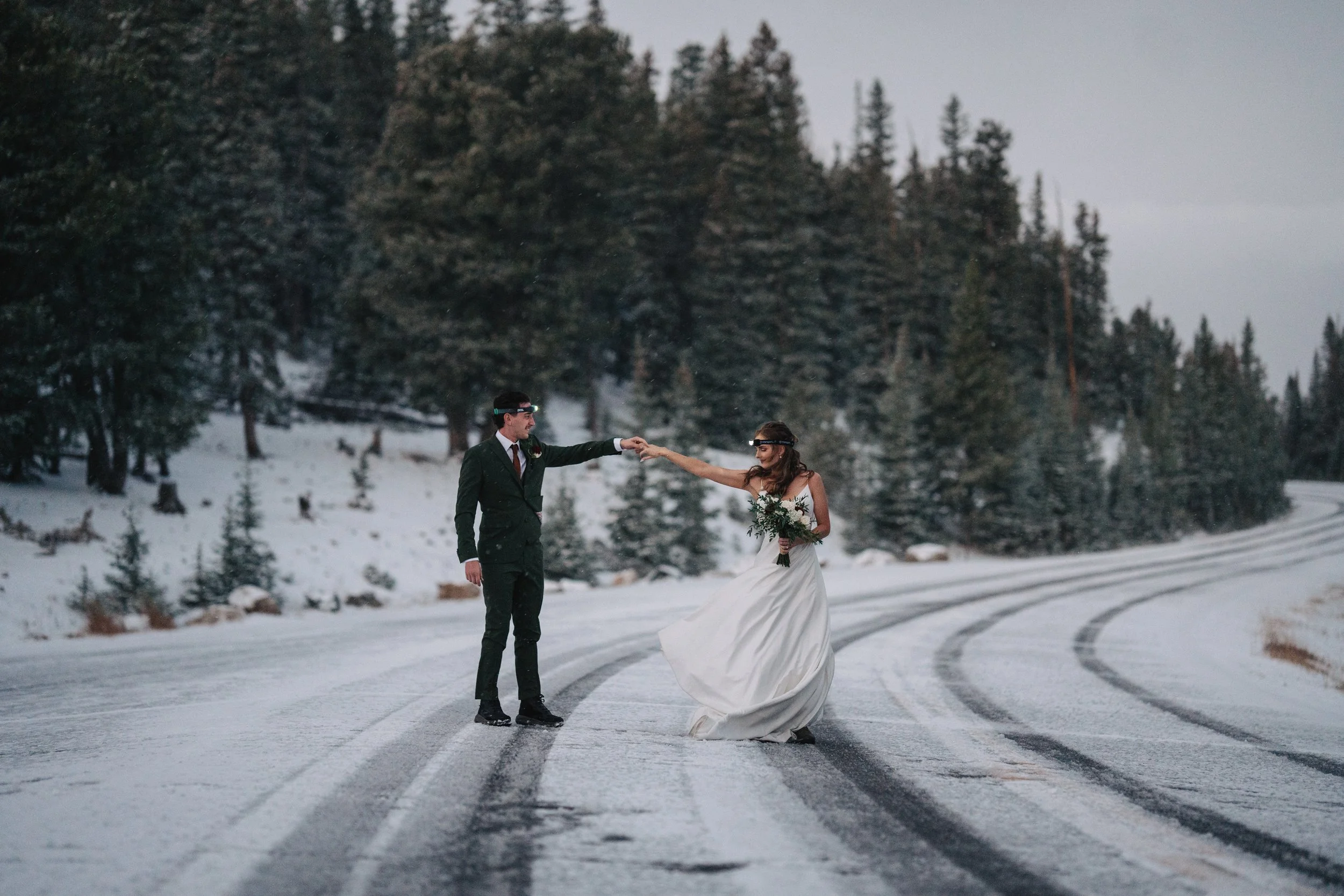 A bride and groom dance on a snow-covered road in a wintery forest. The bride is wearing a white wedding dress and holding a bouquet, the groom is in a dark suit. They are holding hands, and the groom is reaching out to the bride. The surrounding lan