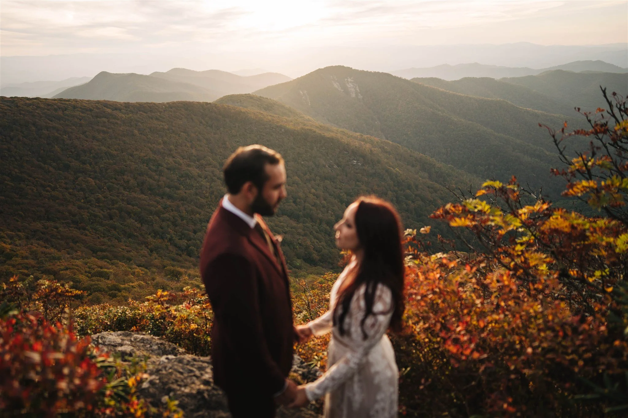 A couple holding hands and looking at each other during sunset in a mountainous, autumn landscape.