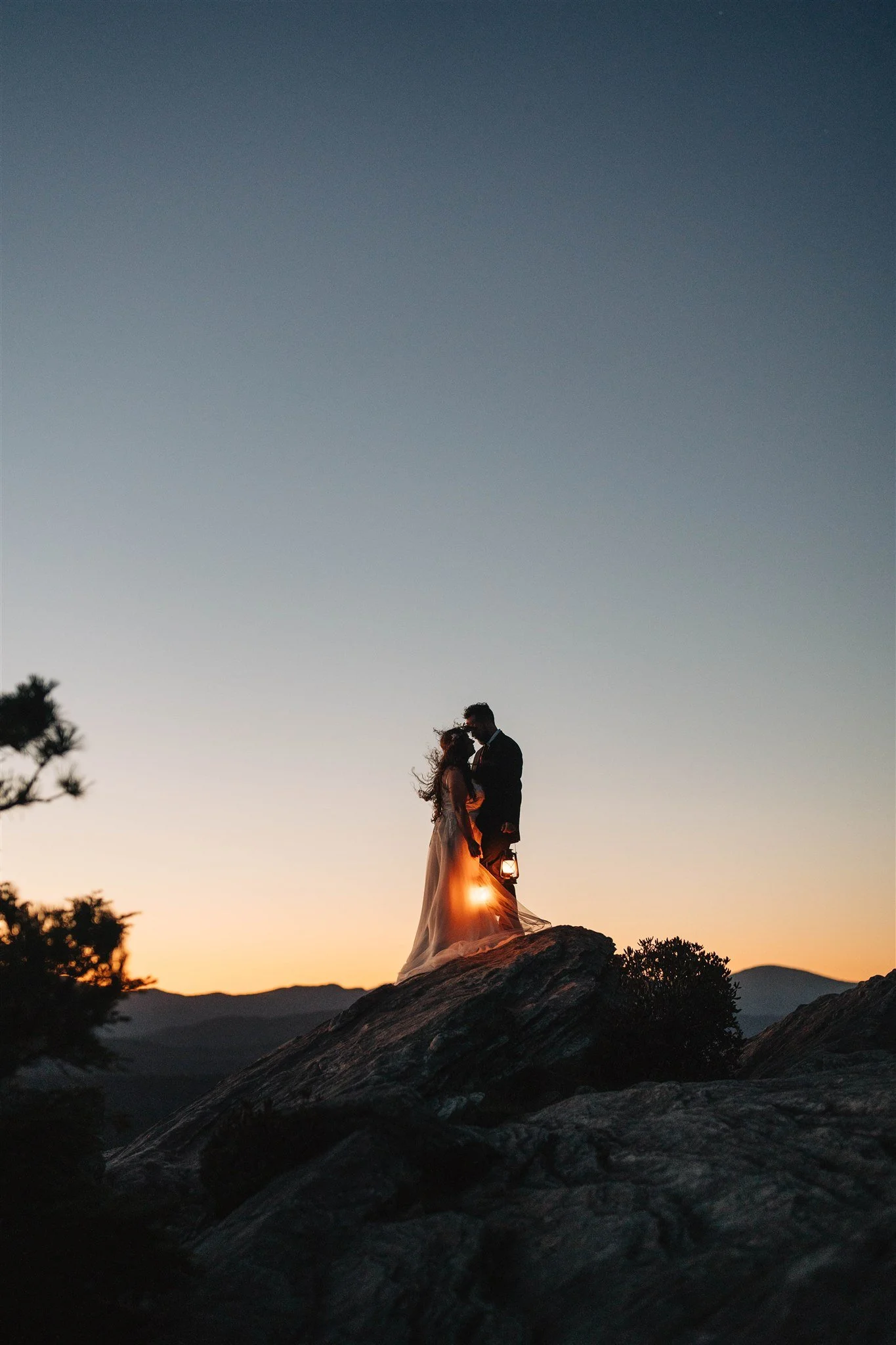 Silhouetted couple standing on a large rock at sunset, with a sunset sky in the background and distant mountains.