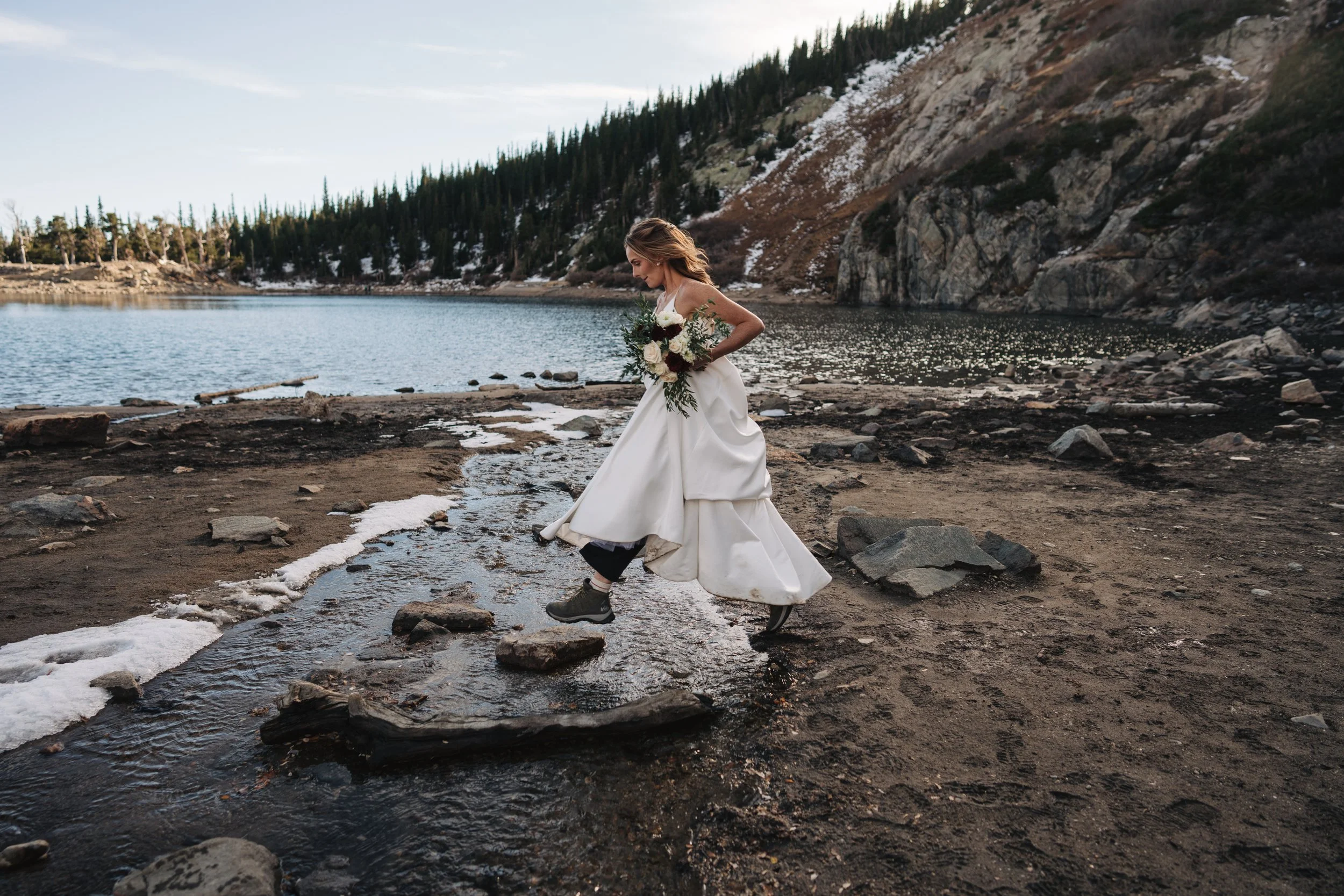A woman in a wedding dress holding a bouquet of flowers, walking on rocks by a lake with mountains and pine trees in the background.