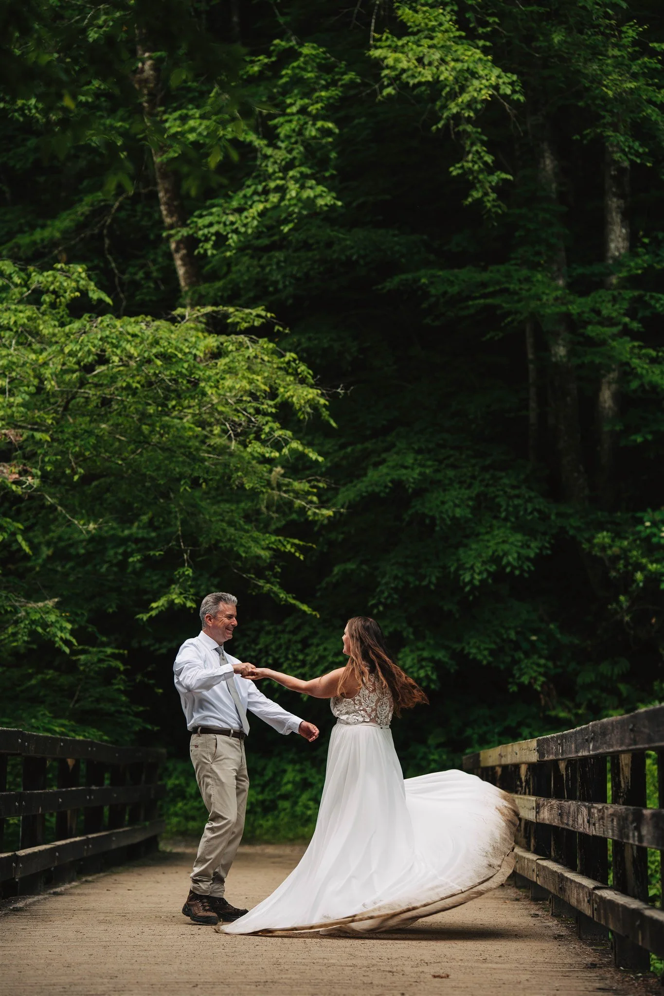 A couple dancing on a wooden bridge surrounded by lush green trees, with the bride wearing a white wedding dress and the groom in a white shirt and khakis.