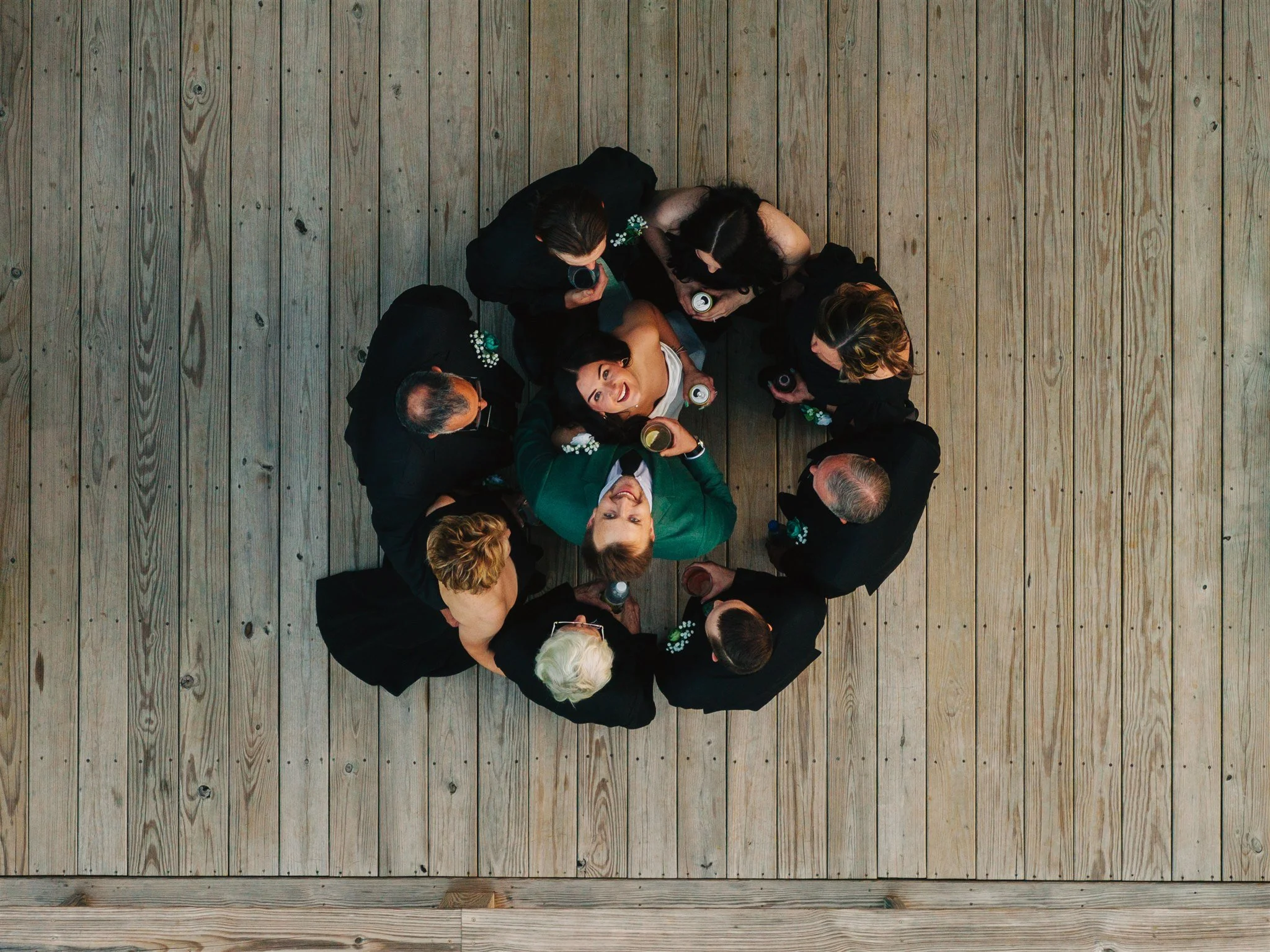 Top-down view of a group of people gathered in a circle, holding drinks, on a wooden floor.
