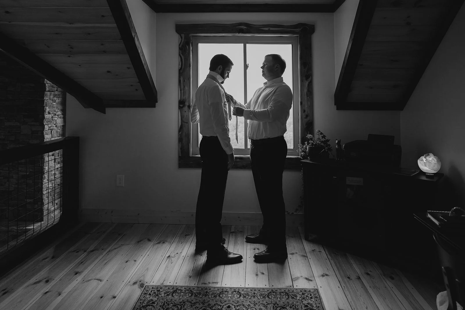 Two men in formal attire standing in front of a window, one helping the other with a tie, in a room with wooden floors and sloped ceiling.