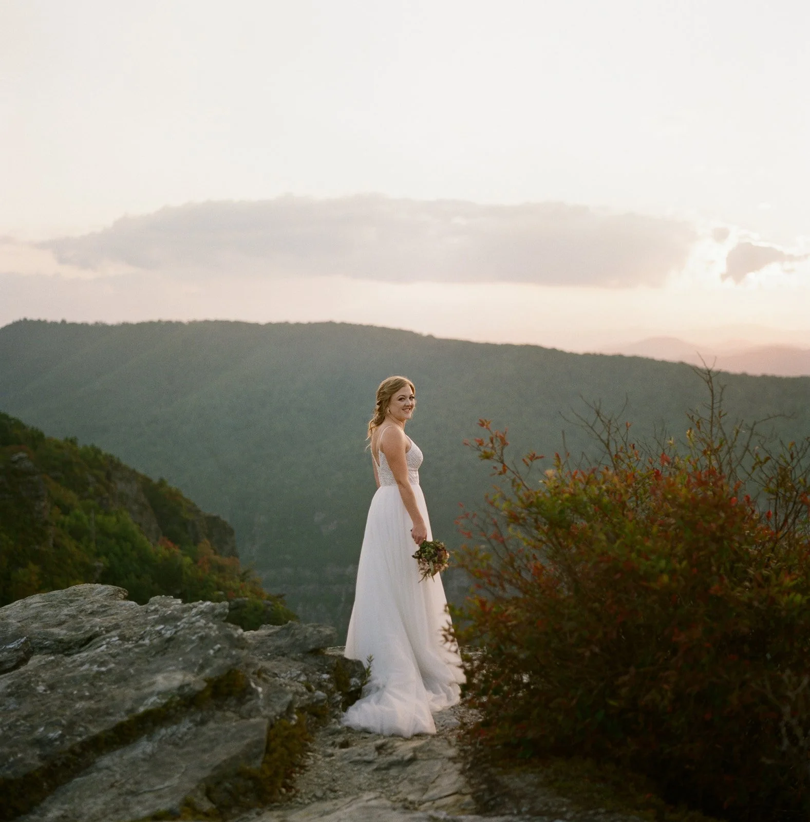 A woman in a white wedding dress holding a bouquet, standing on a rocky ledge with mountains and clouds in the background during sunset.