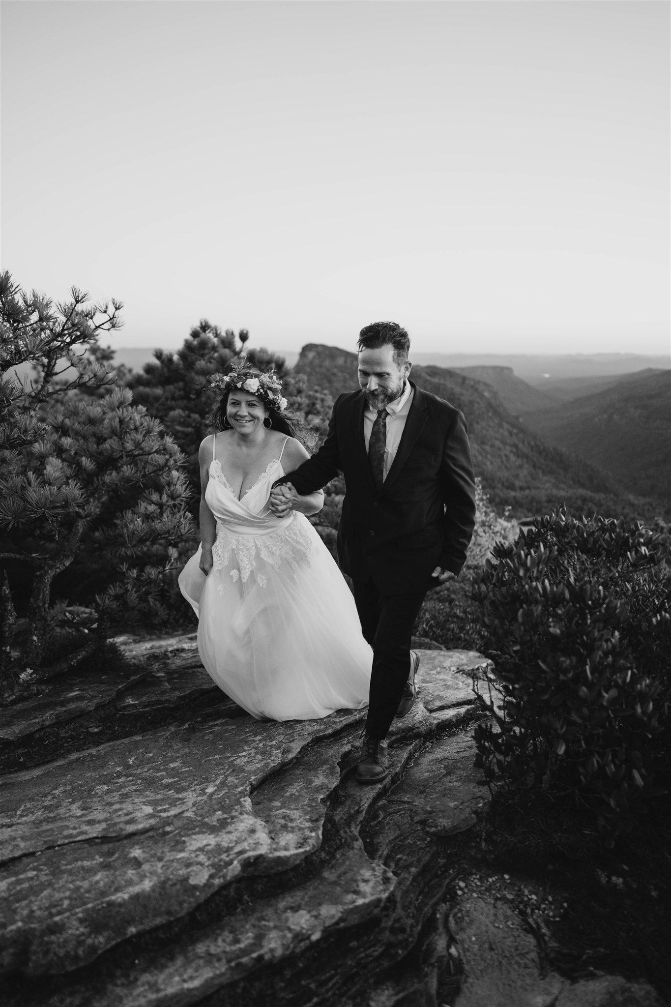 A black-and-white photo of a bride in a wedding dress and a groom in a suit walking hand-in-hand on a rocky outdoor trail with scenic mountains and trees in the background.