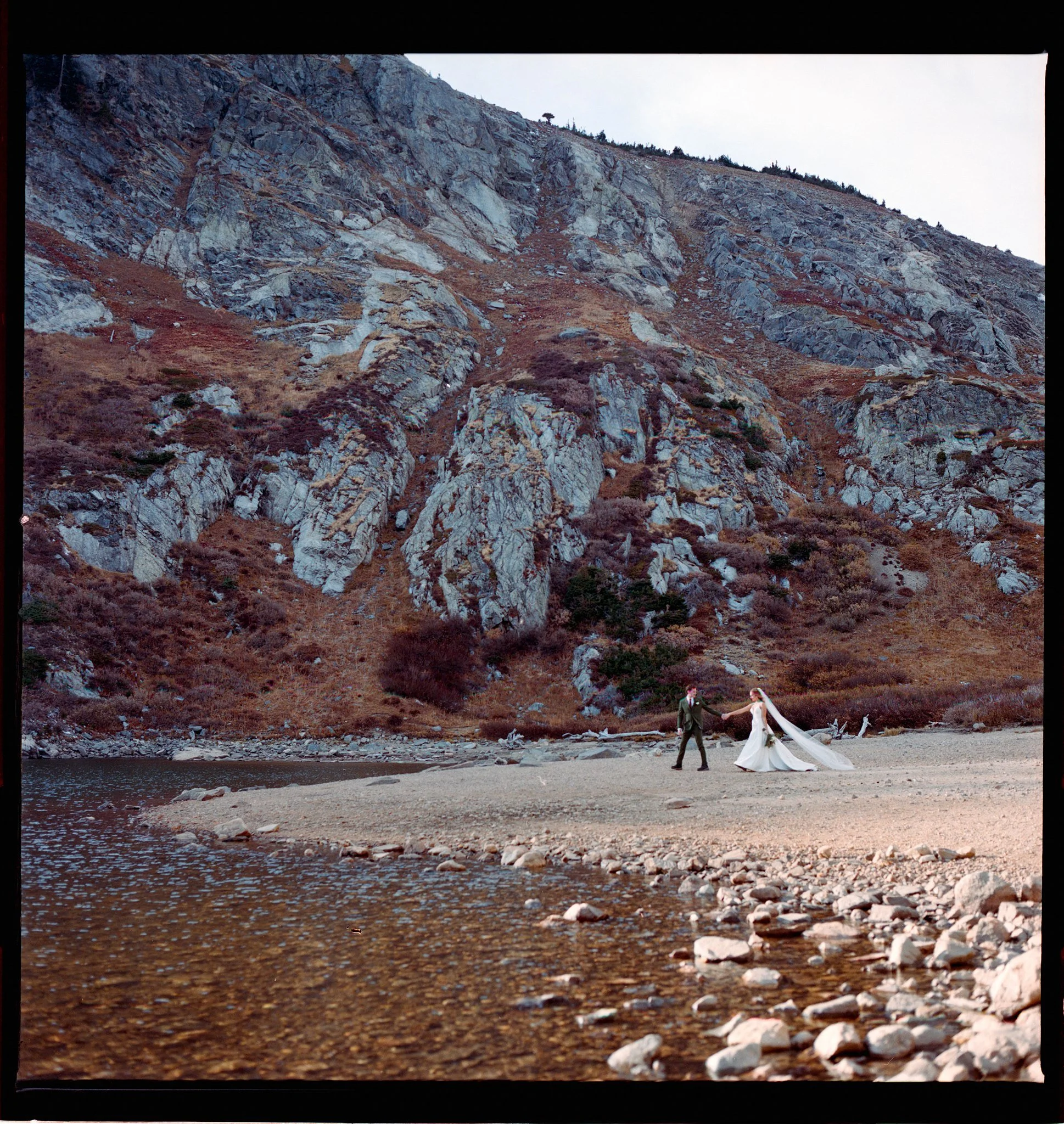 A bride and groom walking hand in hand on a rocky beach with a mountain backdrop.