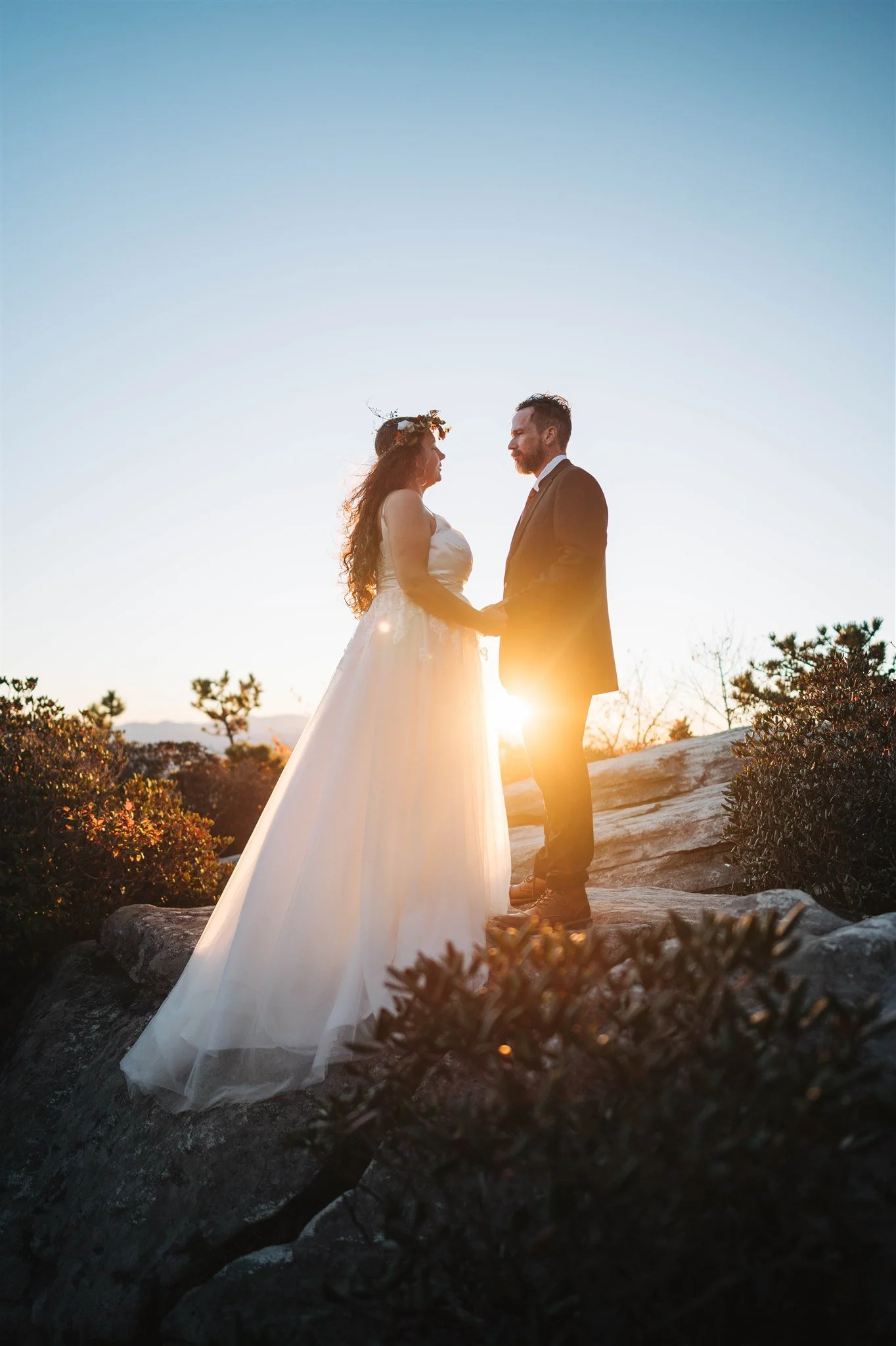 A bride and groom hold hands while facing each other outdoors during sunset, with the sun setting behind them. The bride wears a white wedding dress and a floral crown, and the groom wears a suit. There are rocks and bushes in the foreground and tree
