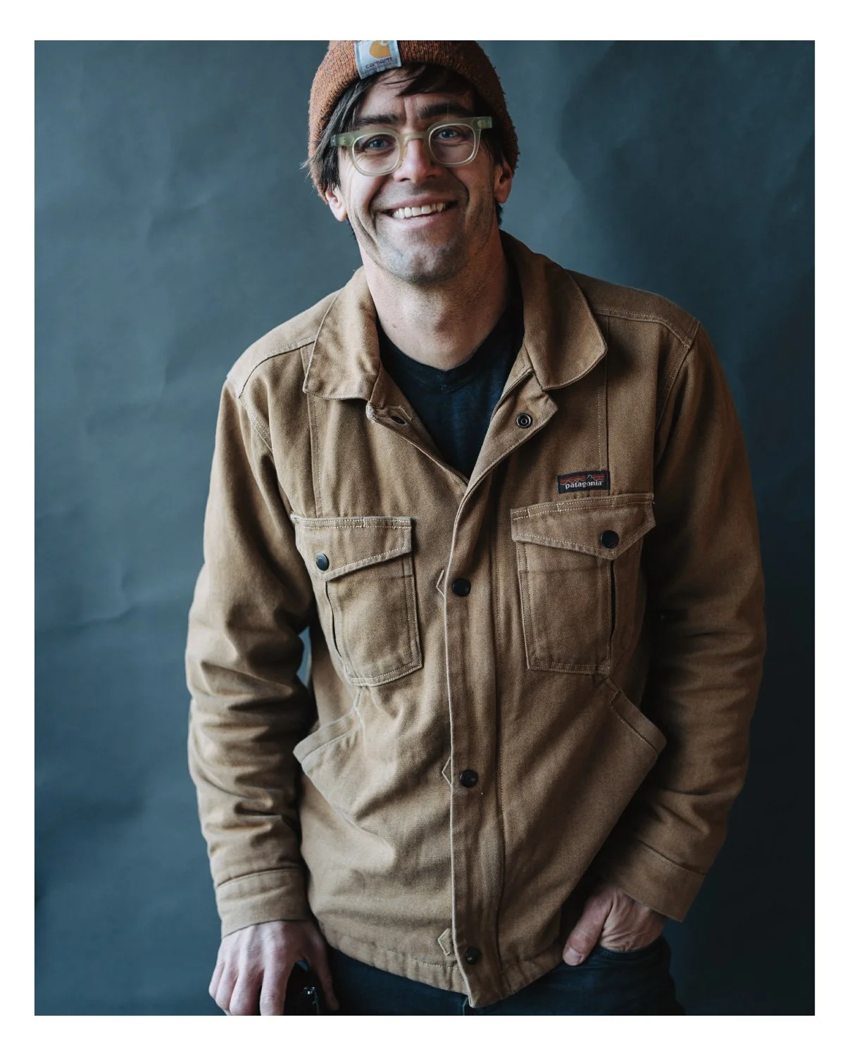 Person wearing brown jacket and beanie, smiling against a dark background
