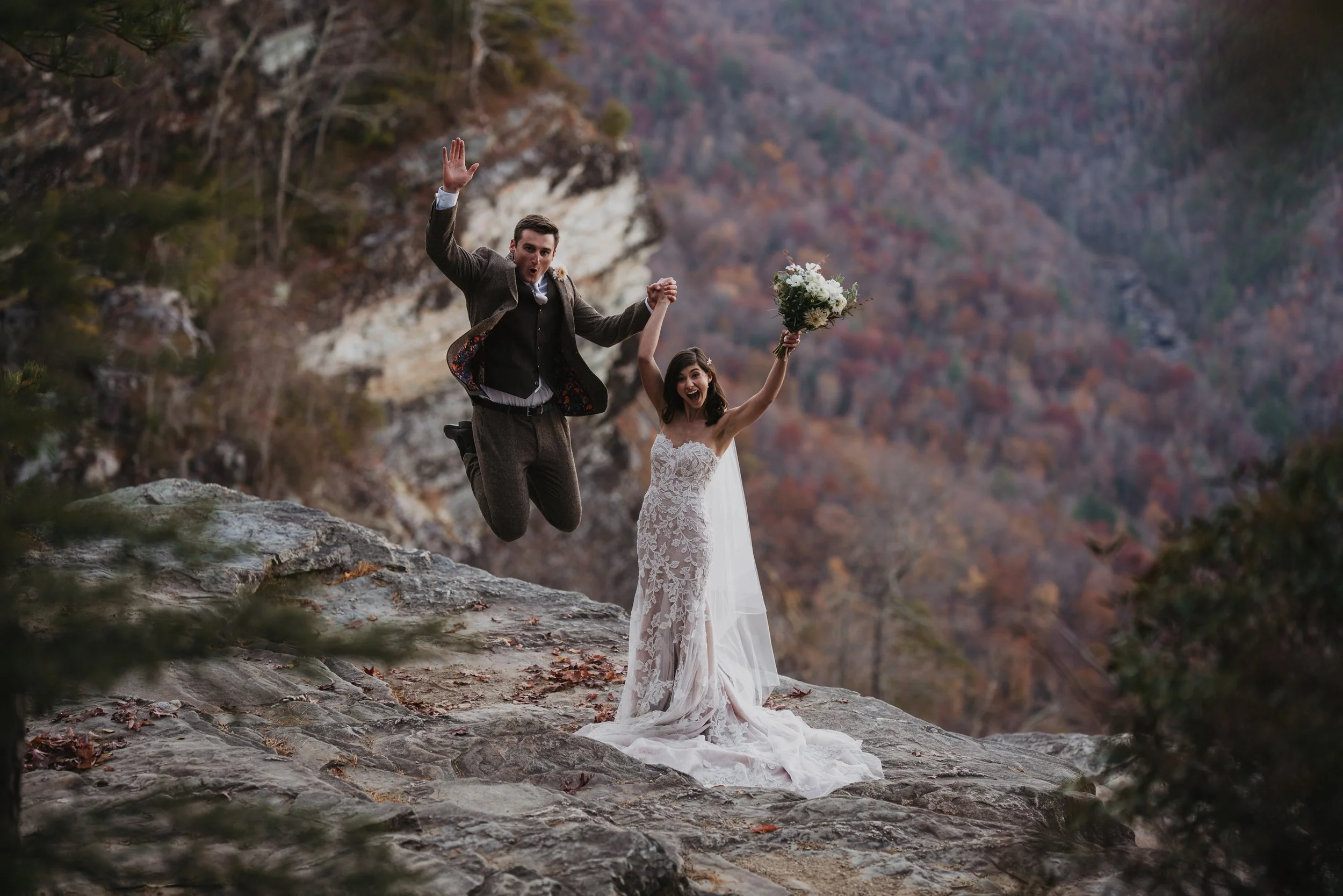 A groom jumping in the air and a bride holding a bouquet outdoors, with a mountainous forest background during fall.