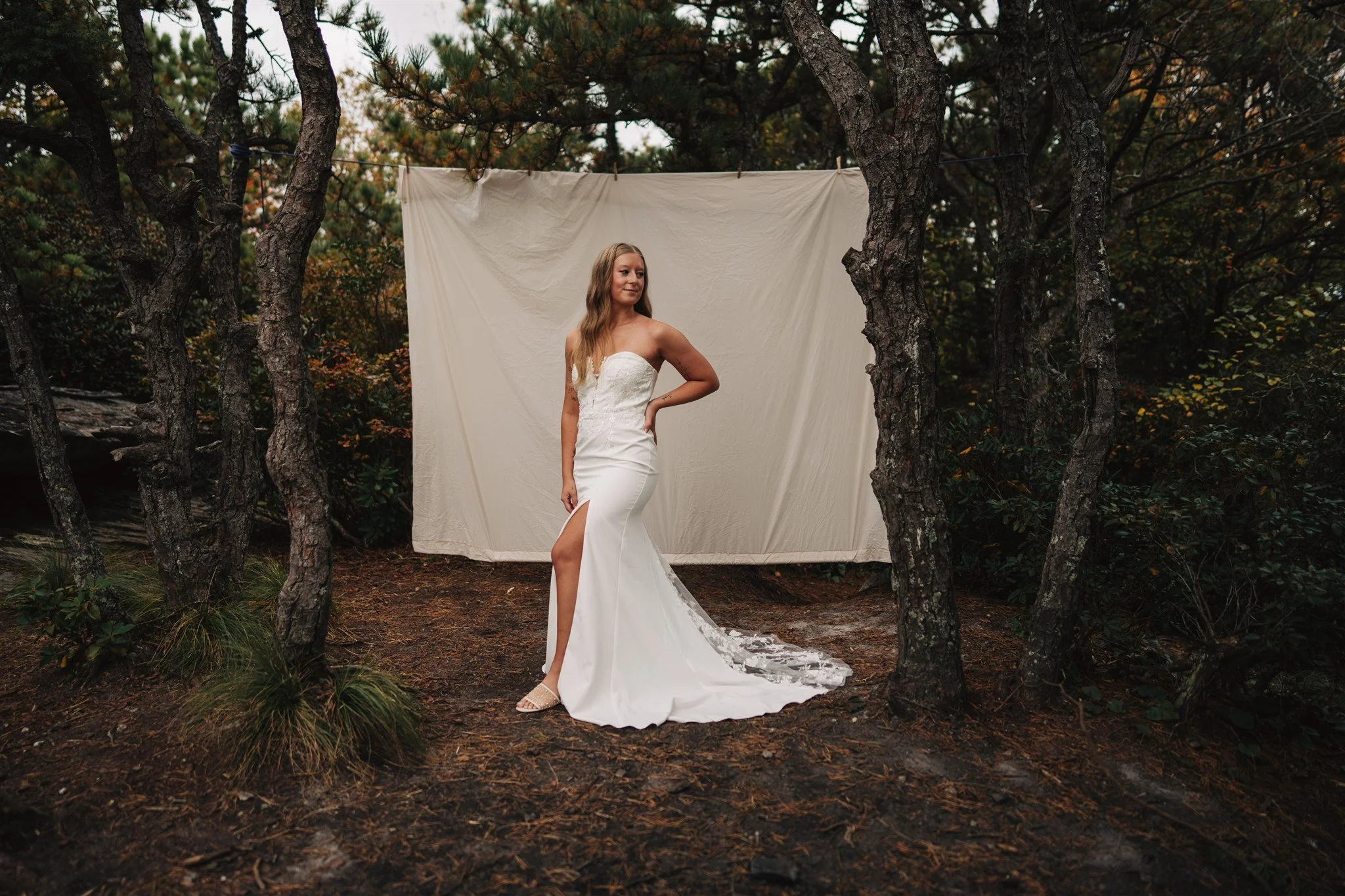 A woman in a strapless white wedding dress with a high slit standing outdoors in a wooded area with pine trees, posing in front of a large white backdrop.