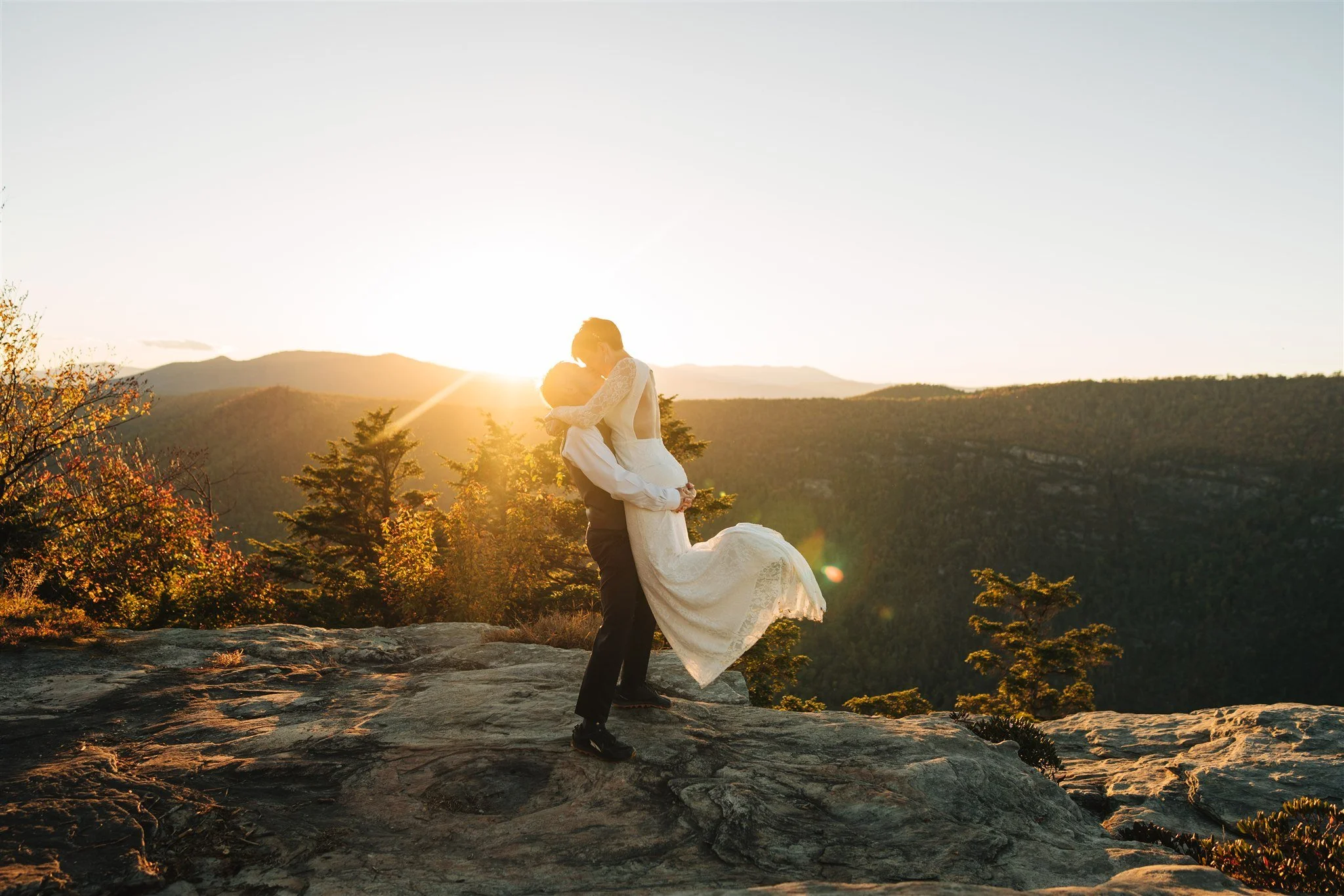 A couple in wedding attire embracing on a rocky hilltop at sunset, with mountains and trees in the background.