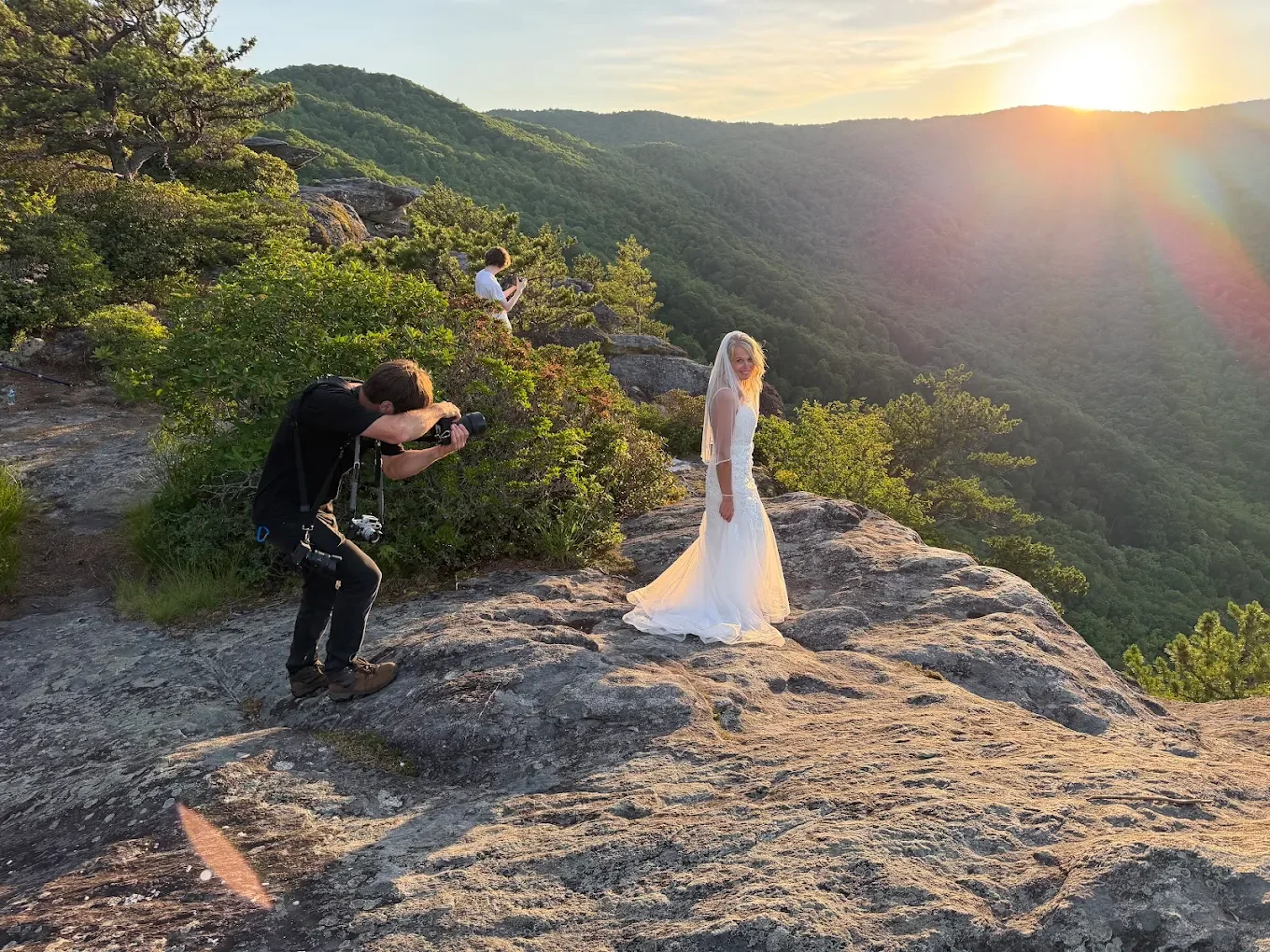 A bride in a white wedding dress standing on a rocky ledge during sunset, while two photographers take pictures of her in a mountainous, wooded area.
