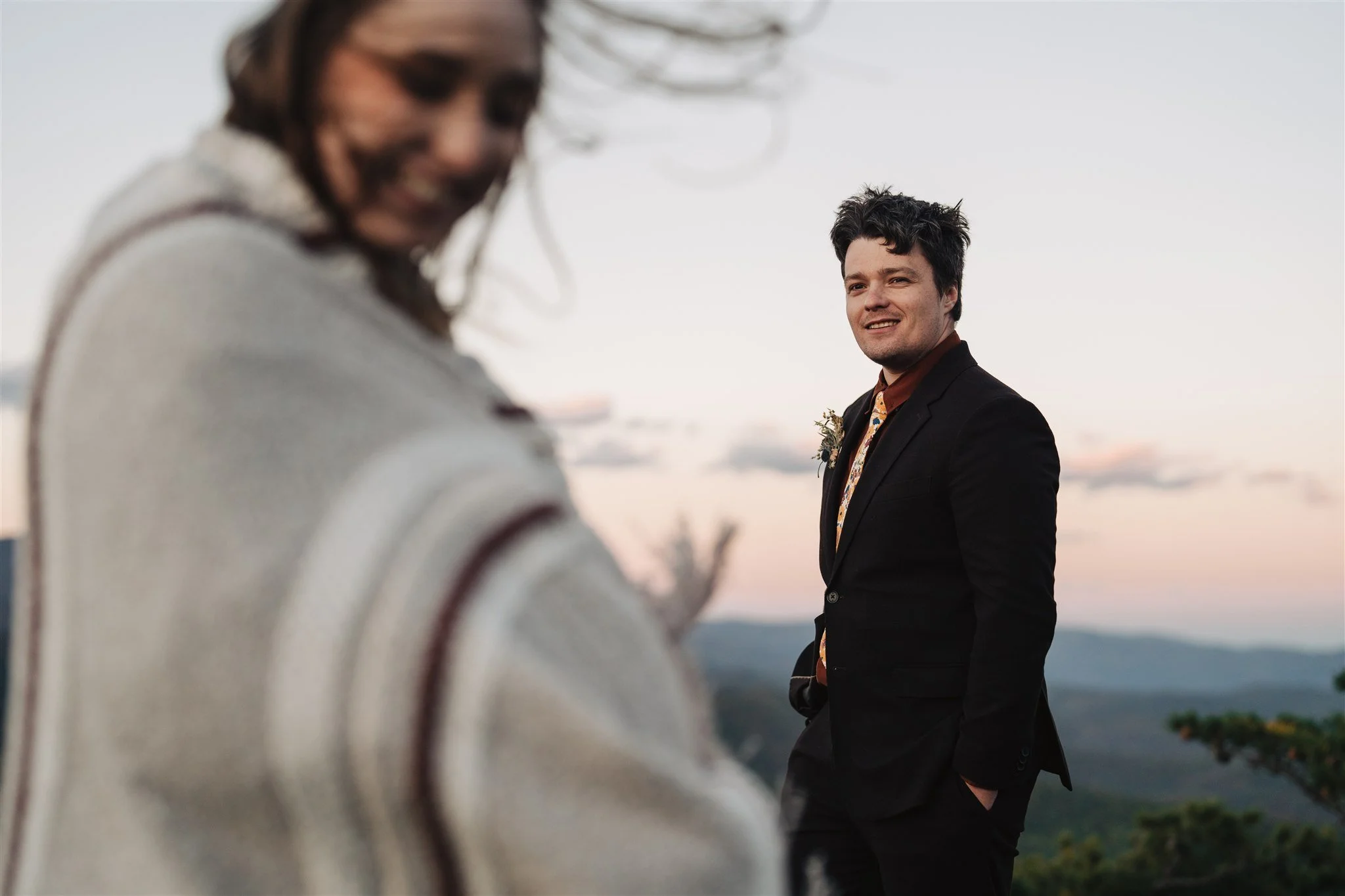 A man in a black suit stands outdoors during sunset, smiling and looking into the distance, with a woman in the foreground off focus, wearing a light-colored sweater.