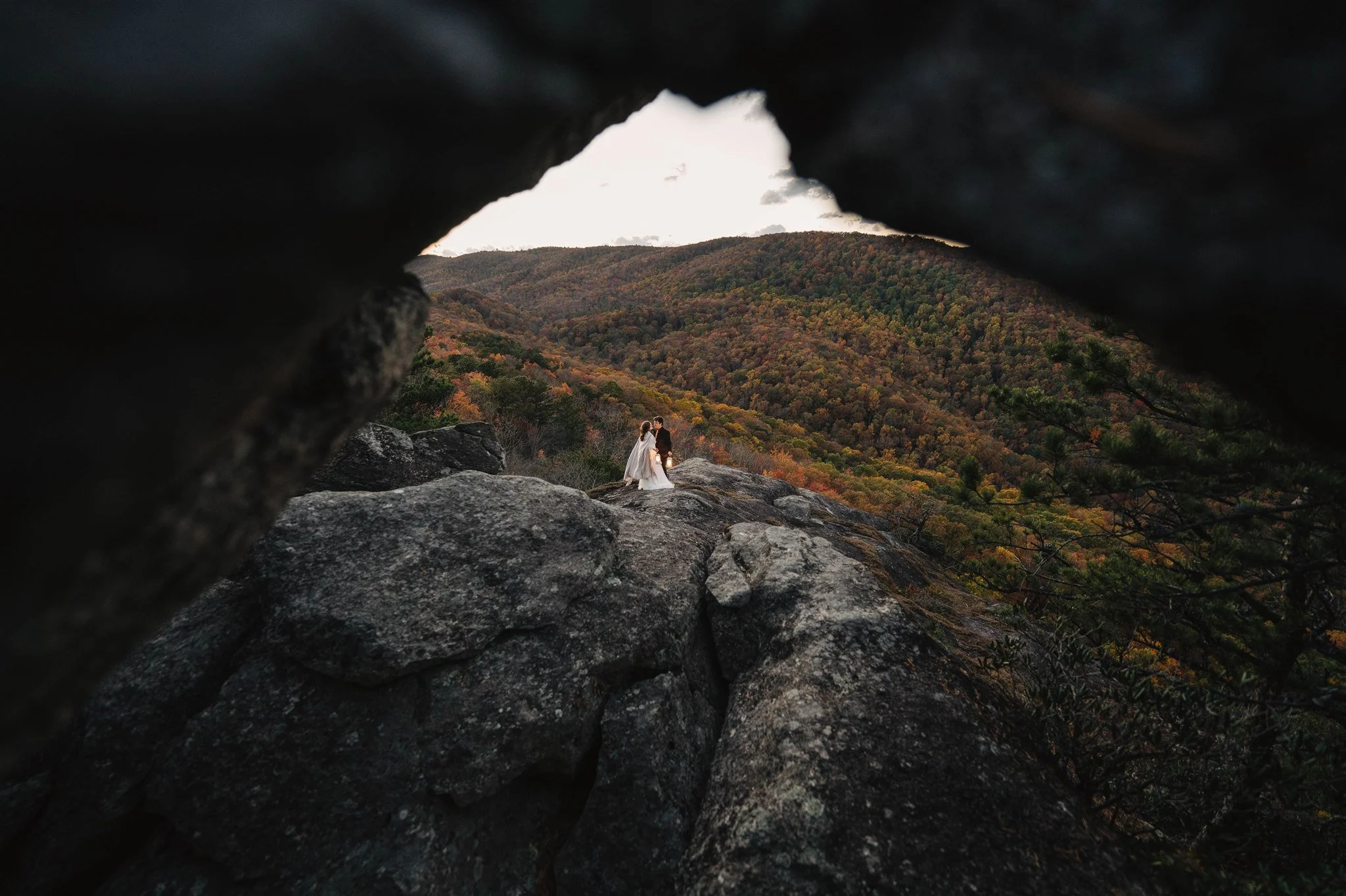A bride and groom holding hands on a large rock, framed by a natural rock opening, with a scenic mountainous landscape in autumn colors in the background.