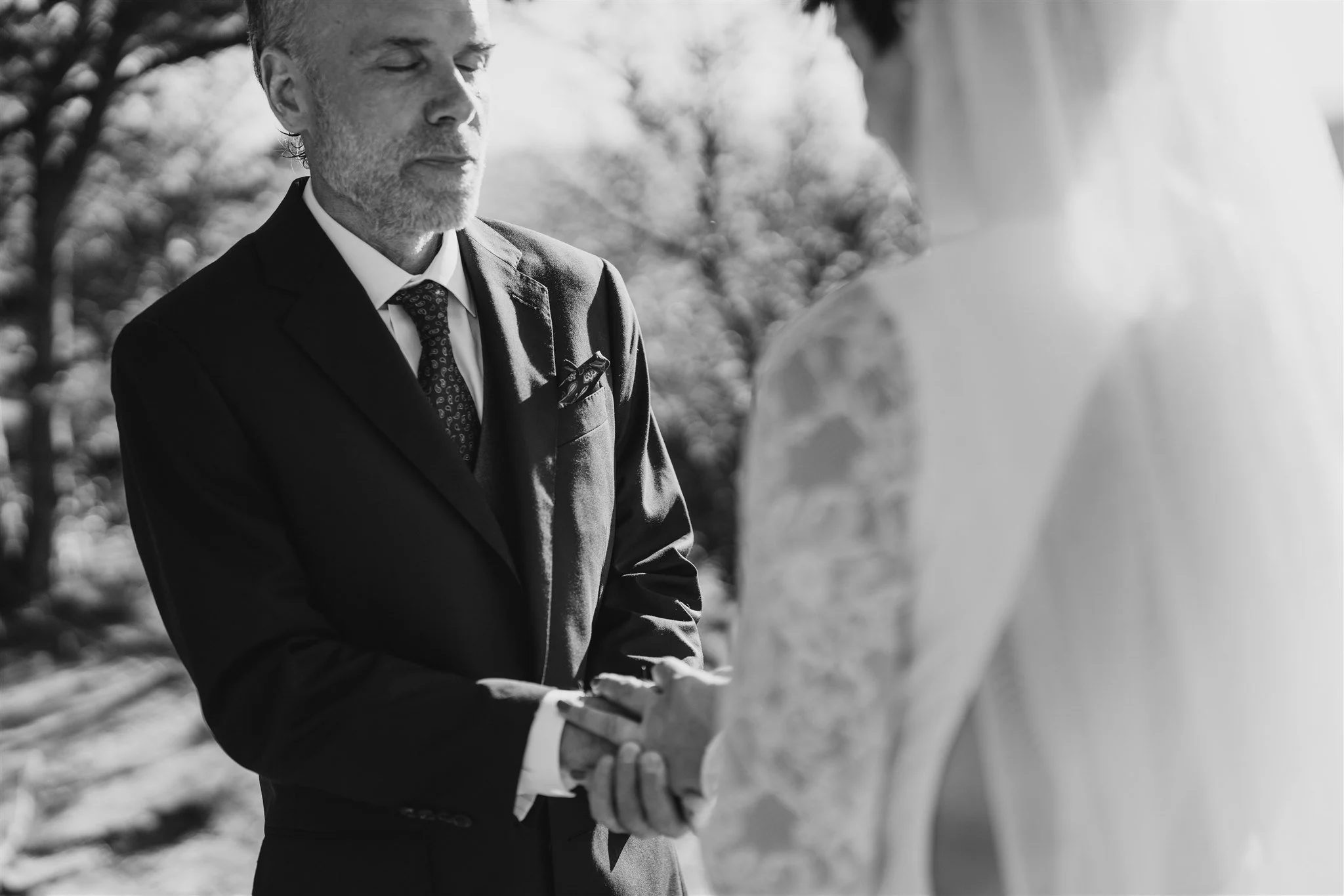 A black and white photo of a man in a suit holding hands with a woman wearing a wedding dress, during a wedding ceremony outdoors.