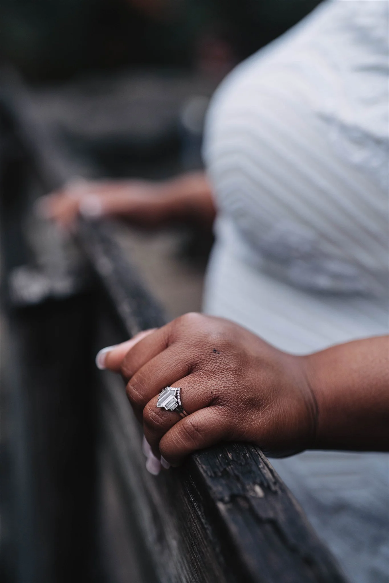 Close-up of a person's hand resting on a wooden railing, wearing a large, elegant diamond ring, with a blurred background of a white striped shirt.