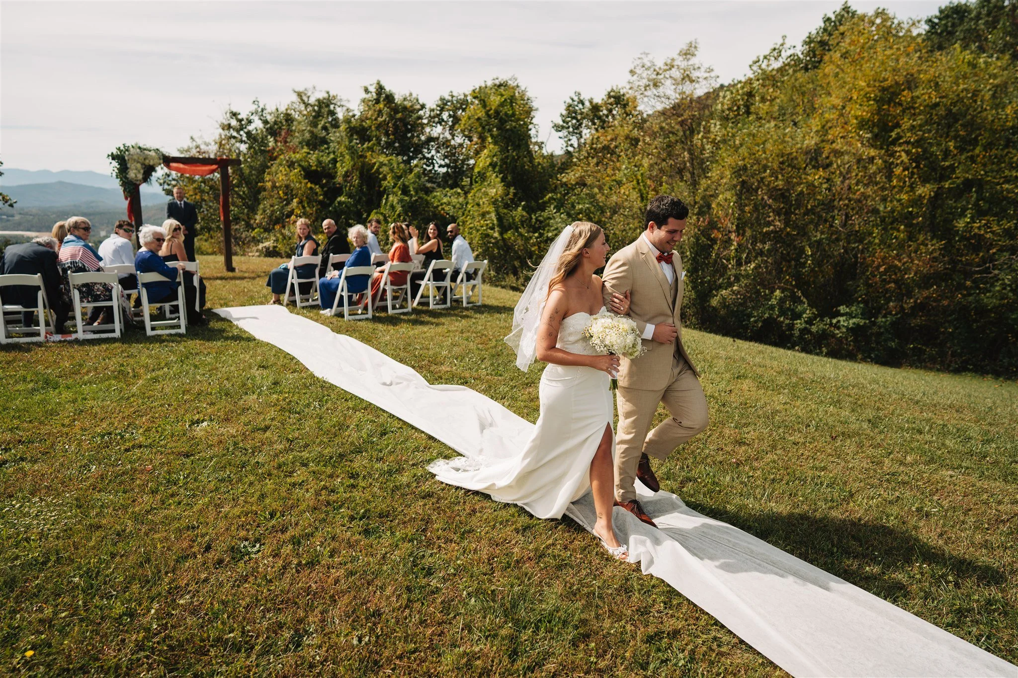 A bride and groom walking arm in arm on a grassy outdoor wedding aisle with guests seated on white chairs in the background.