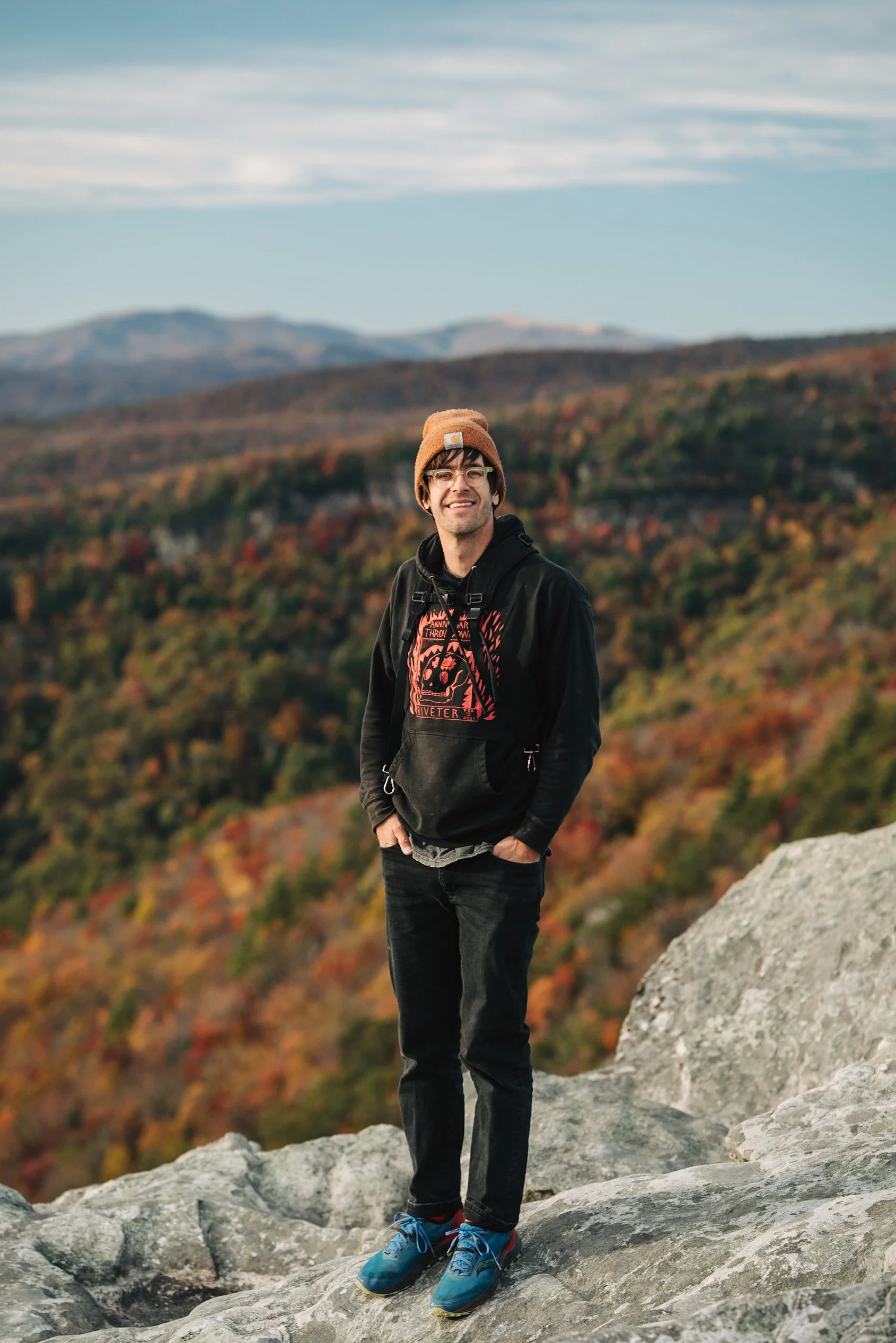 A young man standing on rocky terrain in hiking boots, wearing a black hoodie, black pants, a brown beanie, and glasses, smiling with autumn-colored mountains and sky in the background.