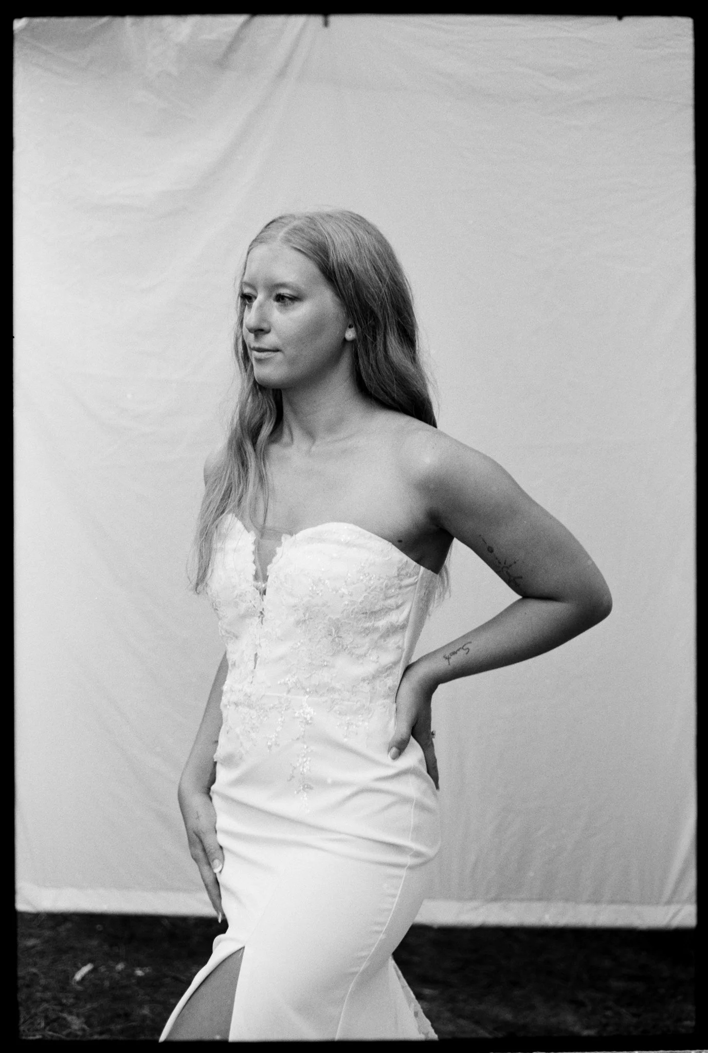 A woman in a wedding dress standing in front of a plain backdrop, looking to her left with her hand on her hip.