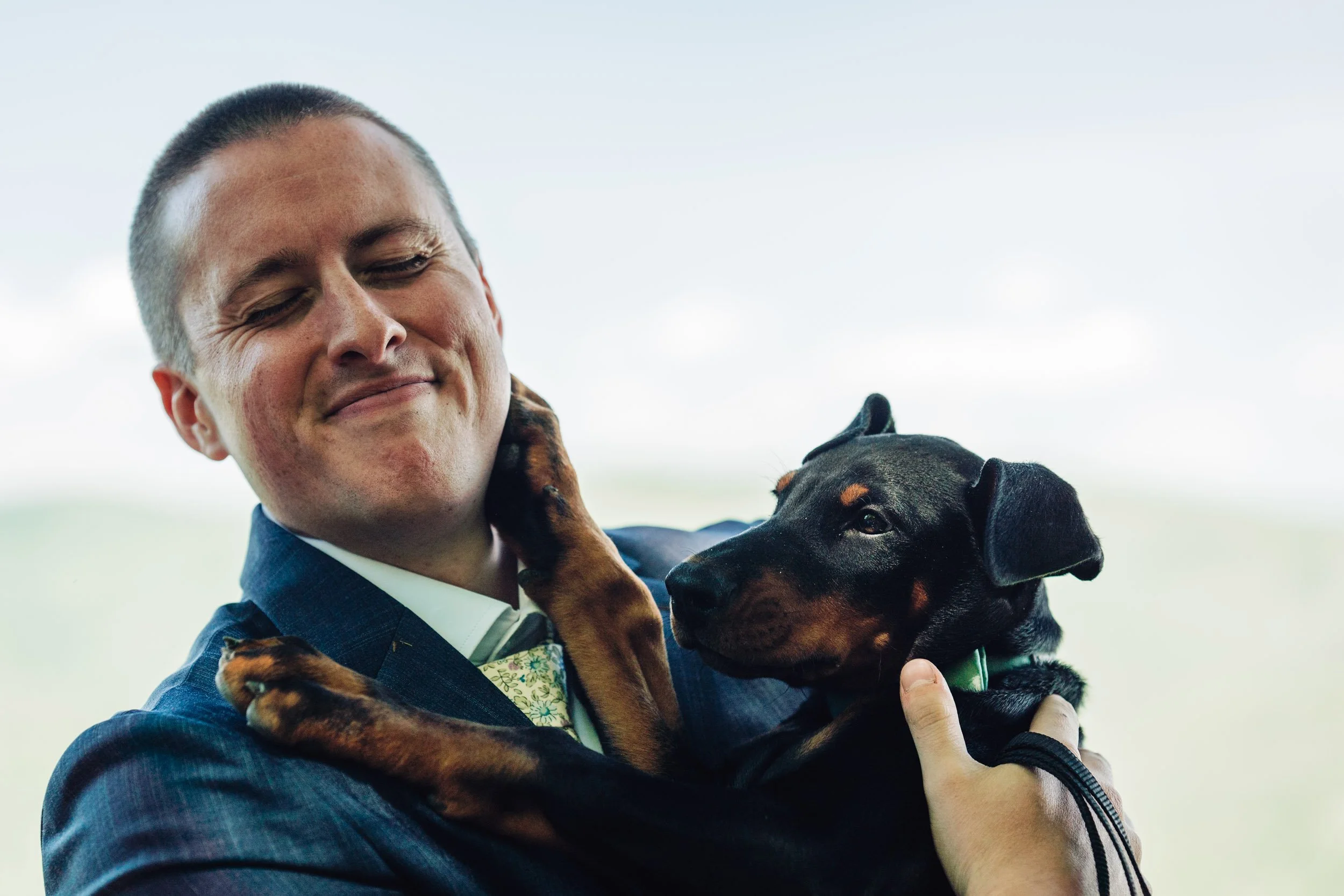 Man in a dark suit holding a black and tan puppy close to his face with a blurred outdoor background.