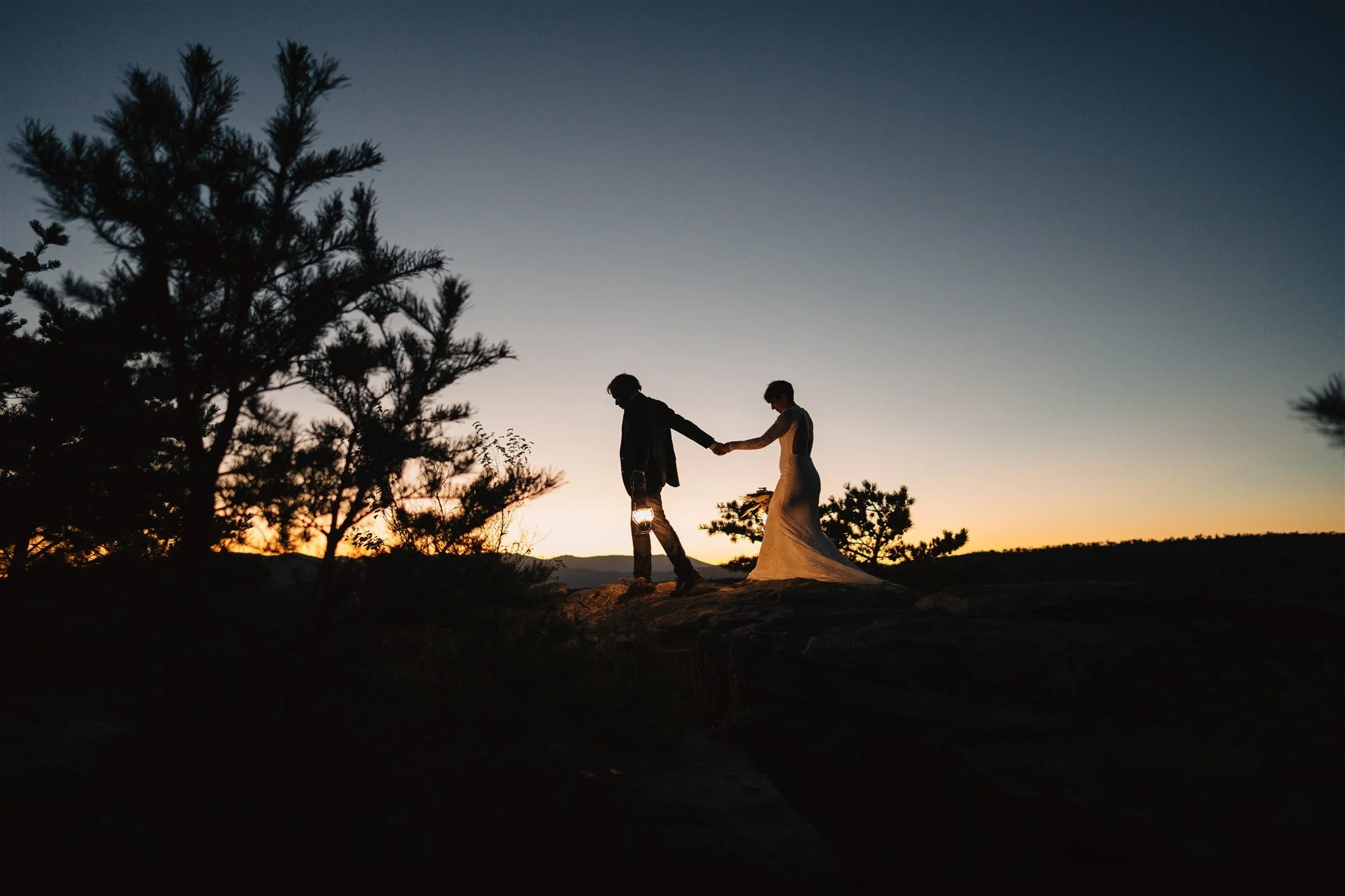 Silhouette of a couple holding hands on a rocky hill at sunset, with trees and a colorful sky in the background.