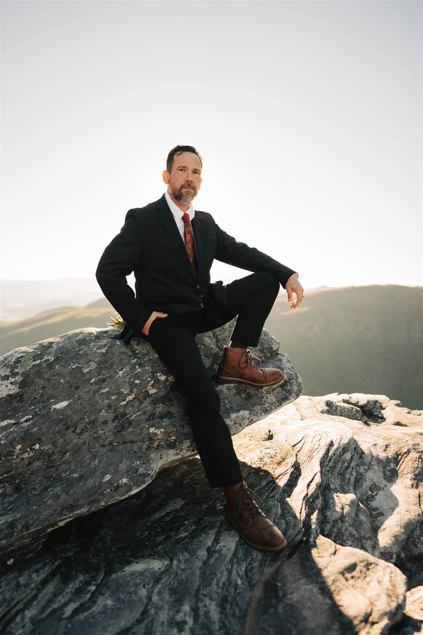 A man in a black suit, white shirt, and brown boots sitting on a large rock outdoors with a scenic landscape and hills in the background.