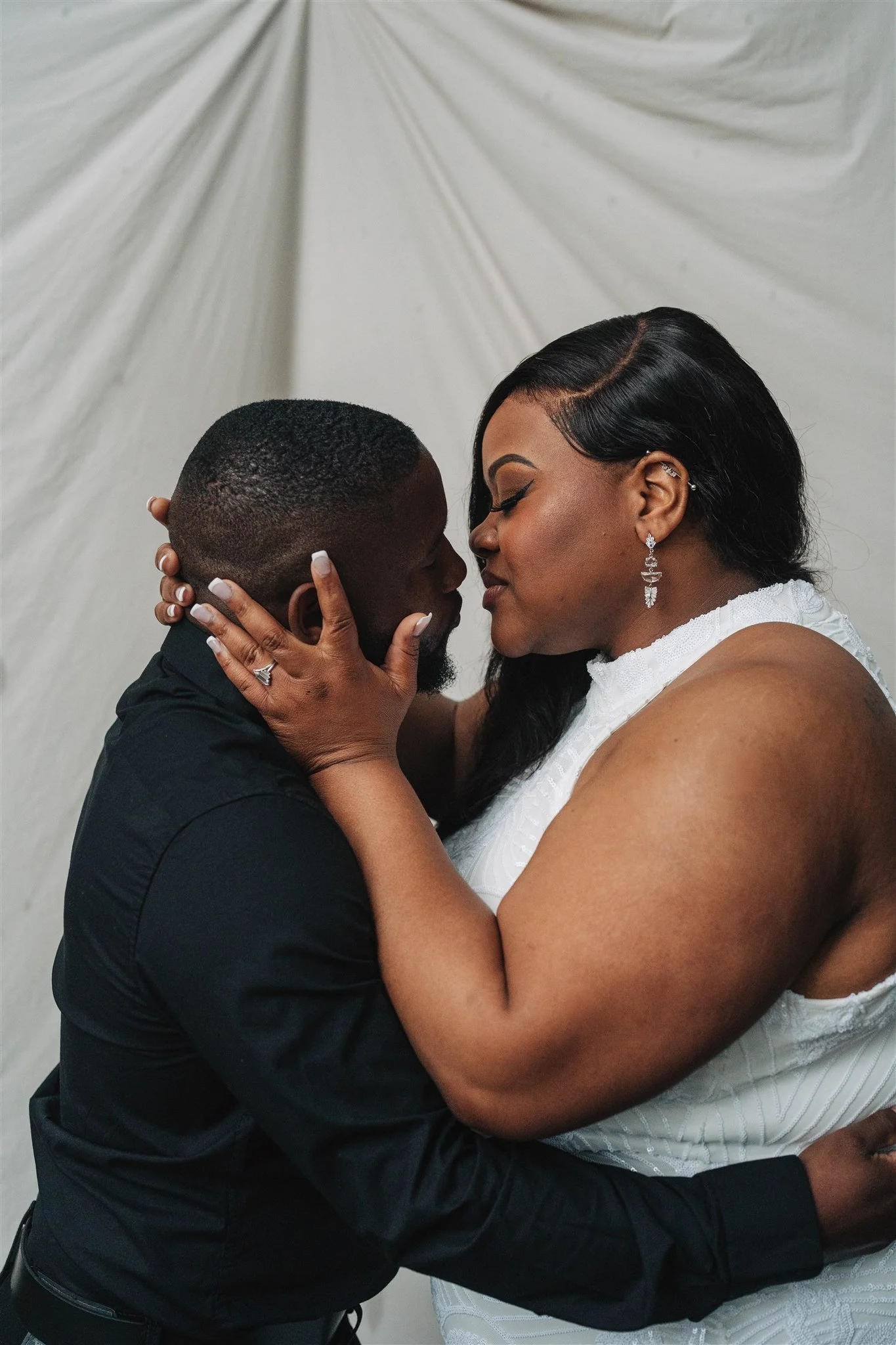 A black couple is embracing, with their foreheads touching, in a romantic moment against a light-colored backdrop.
