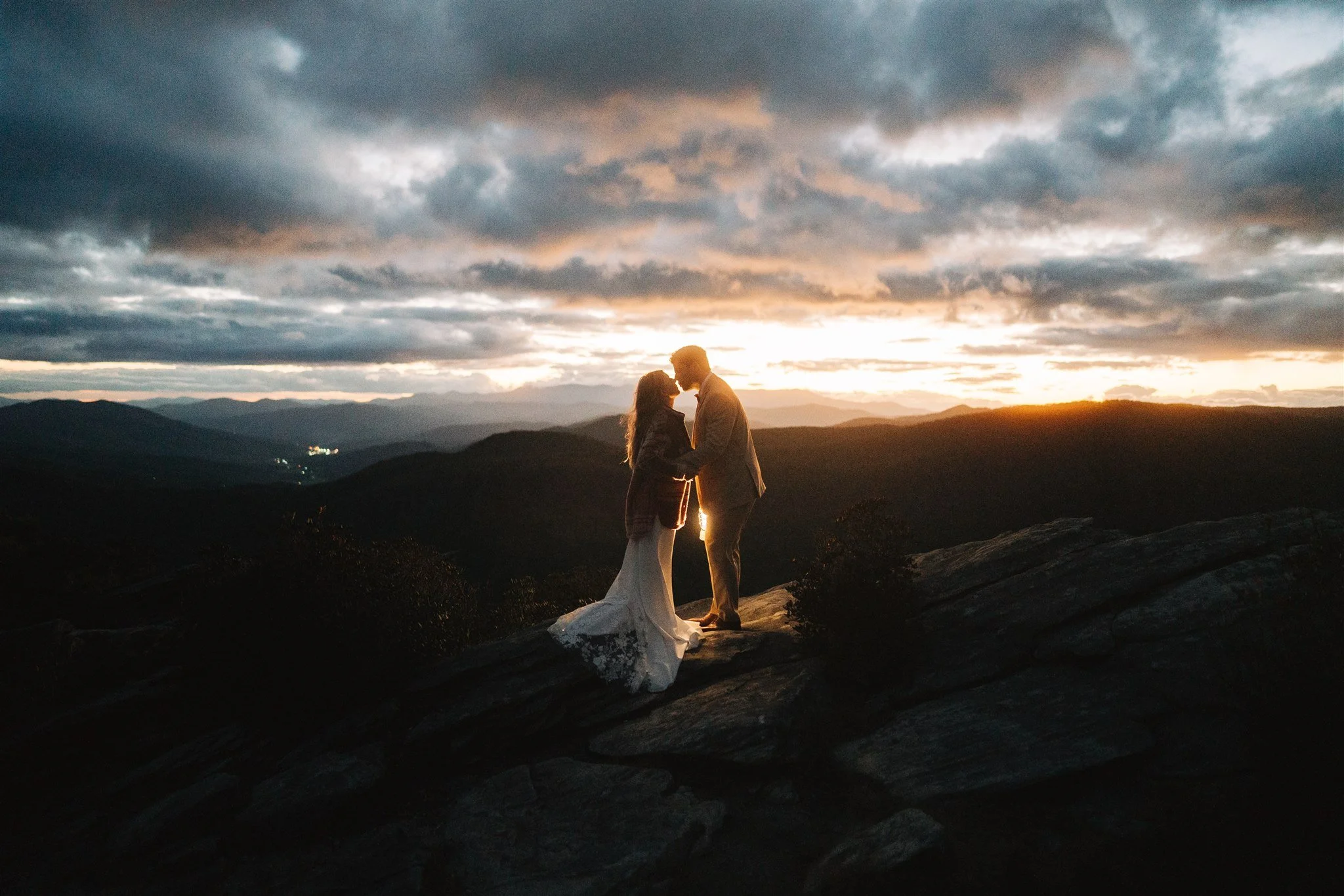 A couple stands on a rocky ledge at sunset, holding hands and leaning in for a kiss with a scenic mountain landscape in the background.