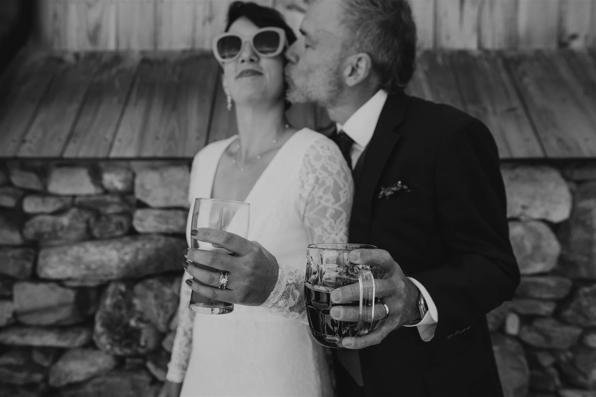 A woman in a white vintage lace wedding dress and sunglasses is holding a glass of beverage, while a man in a suit is kissing her cheek and holding a glass of soda with ice. They are standing in front of a wooden wall with stone accents.