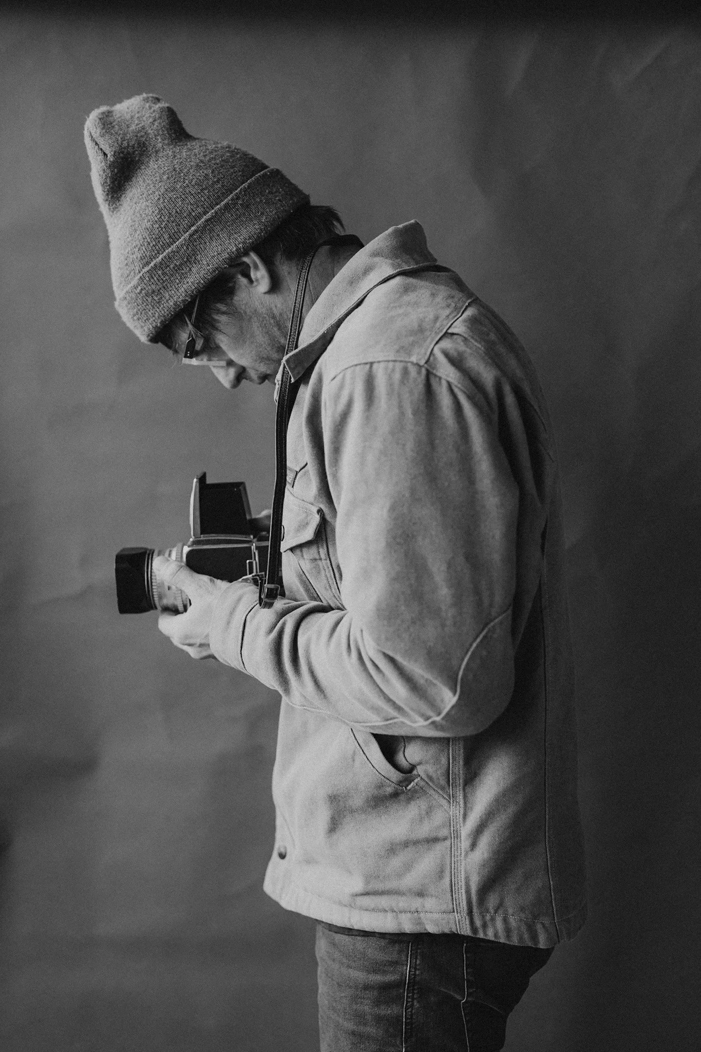 A man in a beanie and glasses looking down and holding a vintage camera.