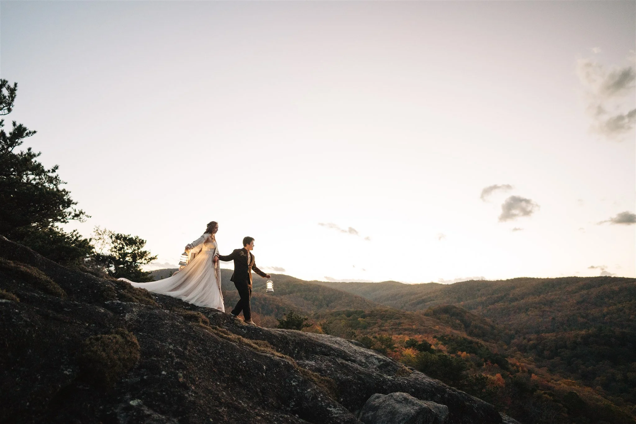 A couple dressed in formal attire walking on a rocky hillside at sunset, with lanterns in their hands, overlooking a mountain landscape and a partly cloudy sky.