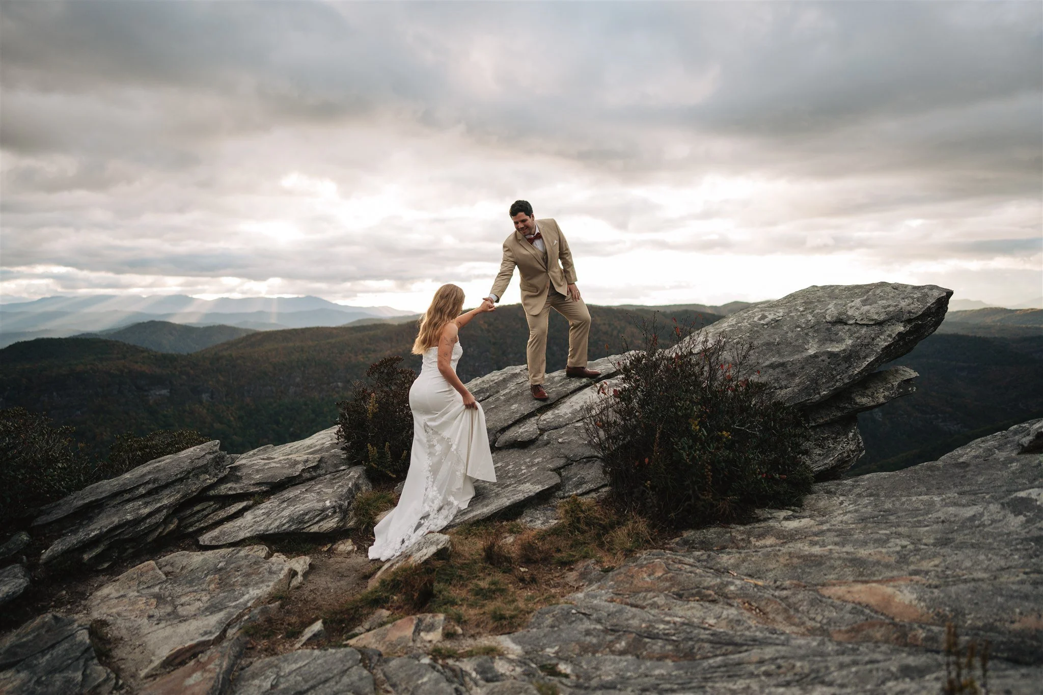A couple, dressed in wedding attire, standing on rocks on a mountain with scenic views of rolling hills and mountains under a cloudy sky. The groom is helping the bride climb up a rock.
