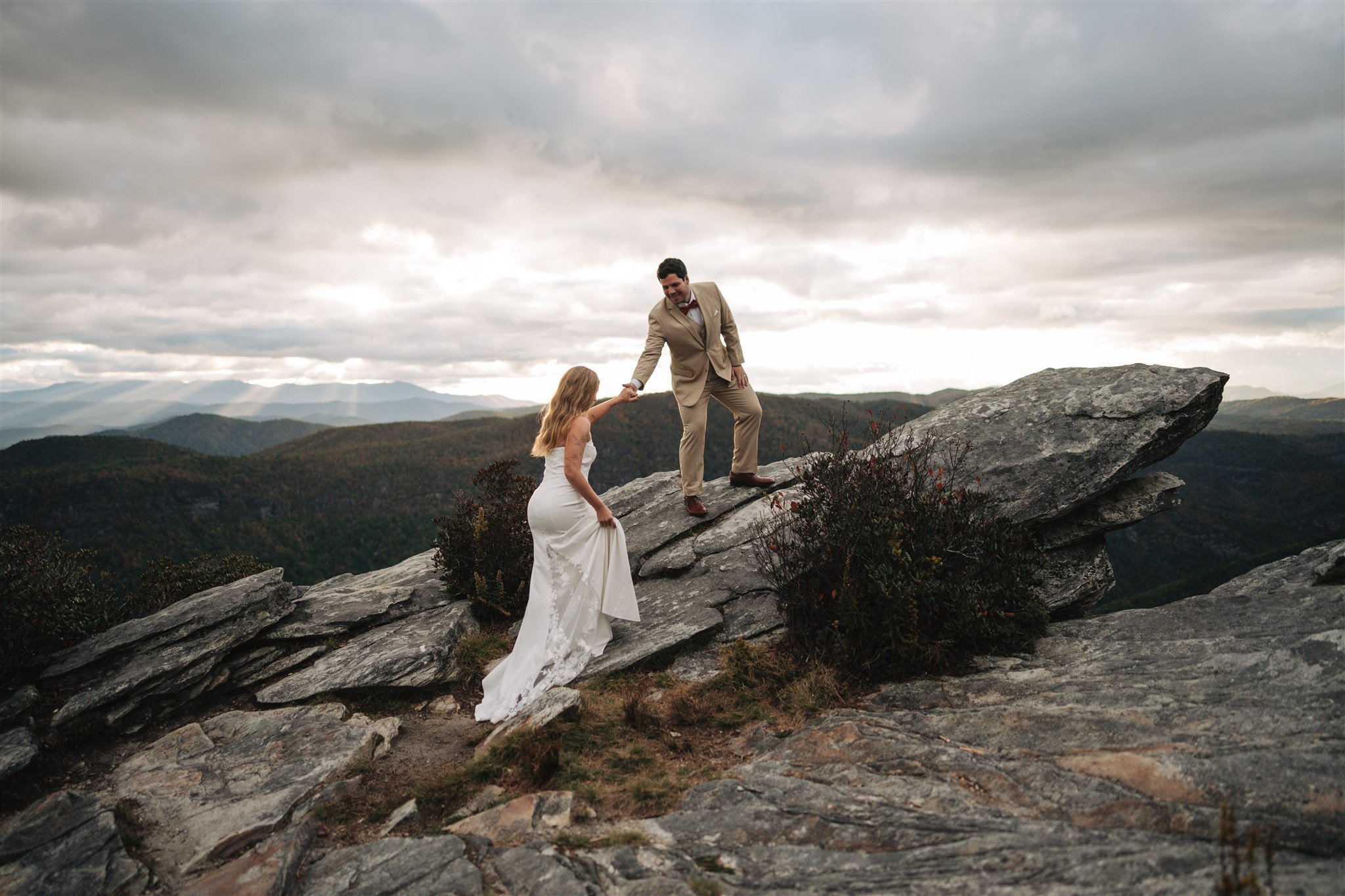 A bride and groom on a rocky mountain ledge, with the groom helping the bride up as they enjoy scenic mountain views under a cloudy sky.