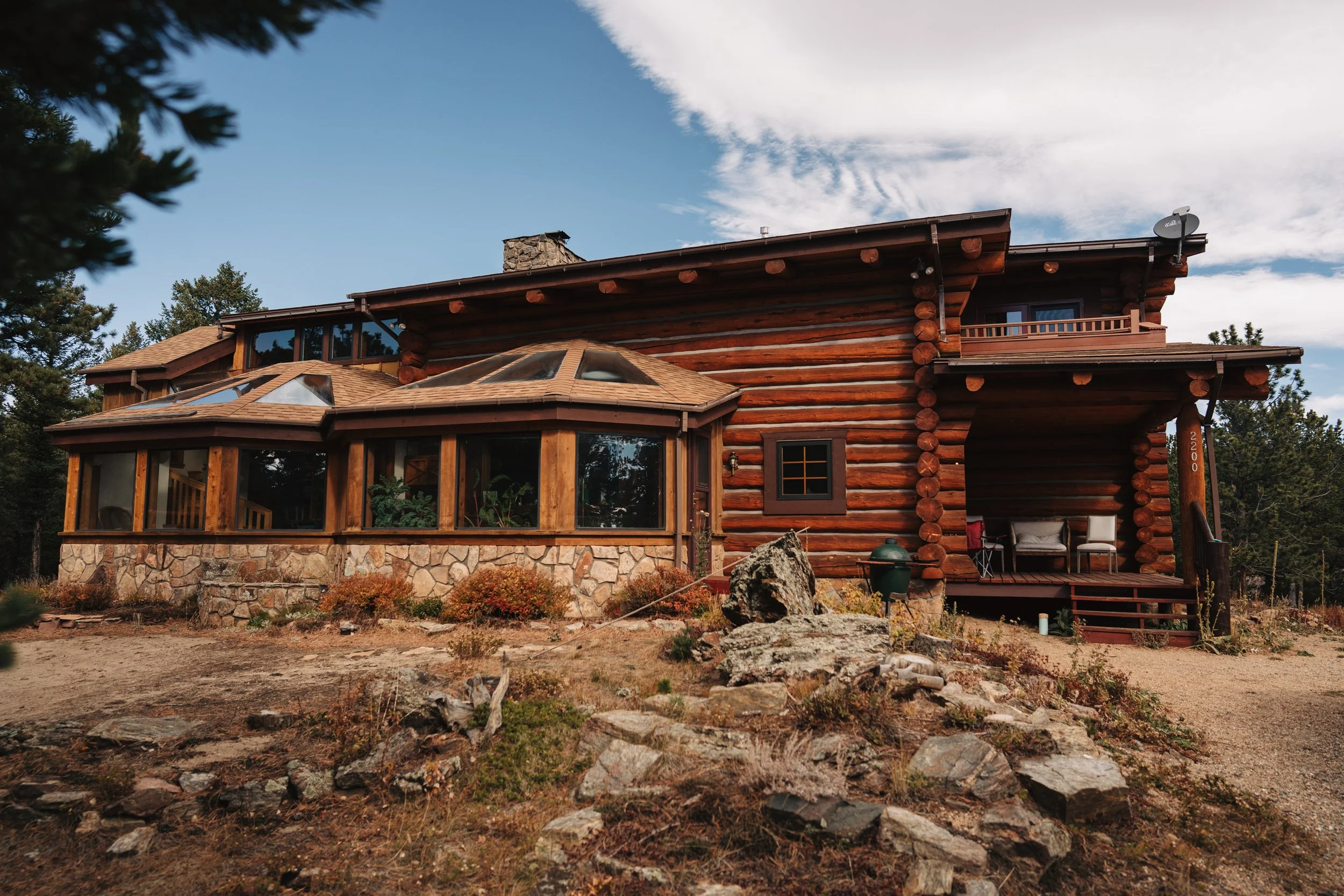 A rustic log cabin with stone foundation surrounded by trees and rocks, featuring large windows and a small porch with seating, under a clear blue sky.